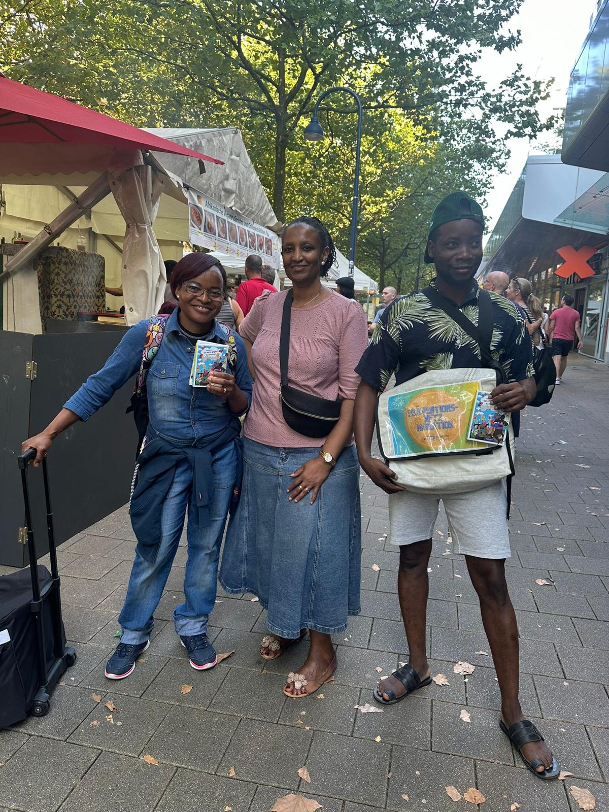 Drei Freunde posieren lächelnd auf einer Marktstraße mit Zelten und Bäumen im Hintergrund; links Brillenträgerin, Mitte rosa