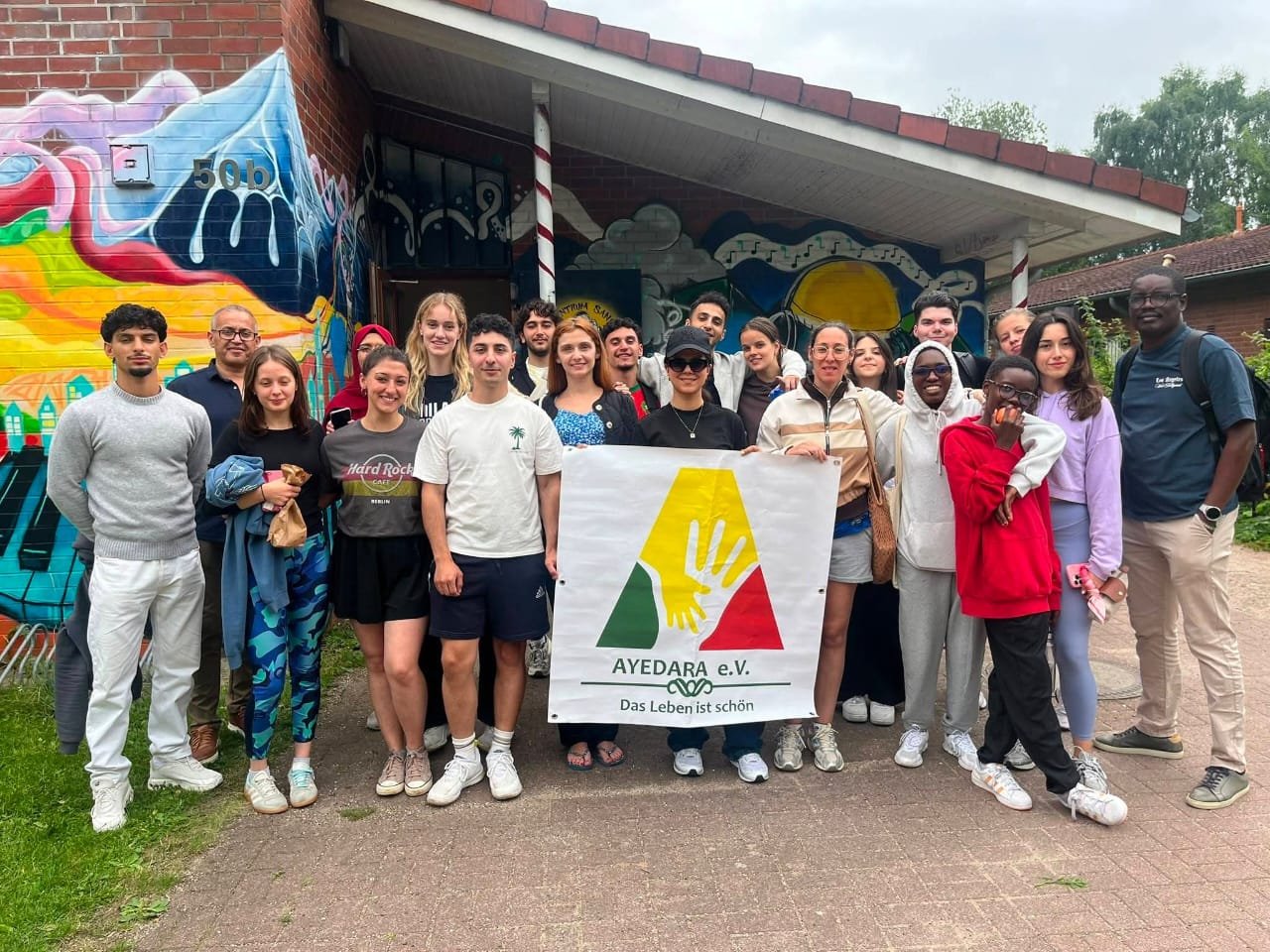 Gruppe junger Menschen posiert vor einer bunten Graffiti-Wand und hält ein Banner von AYEDARA e.V. mit gelber Hand.