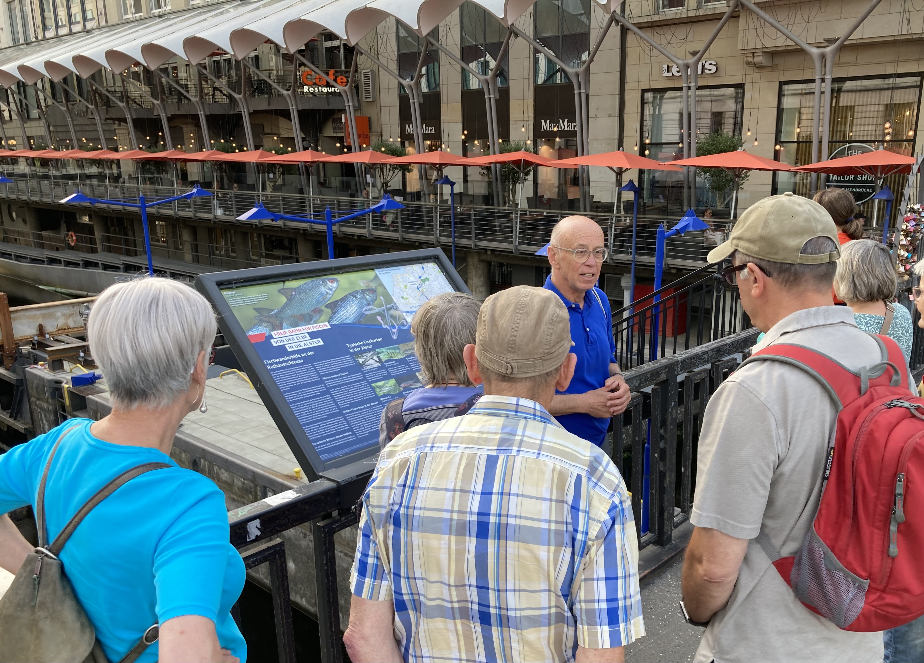 Gruppe von Touristen vor Infotafel, rote Sonnenschirme und moderne Uferpromenade am Kanal.