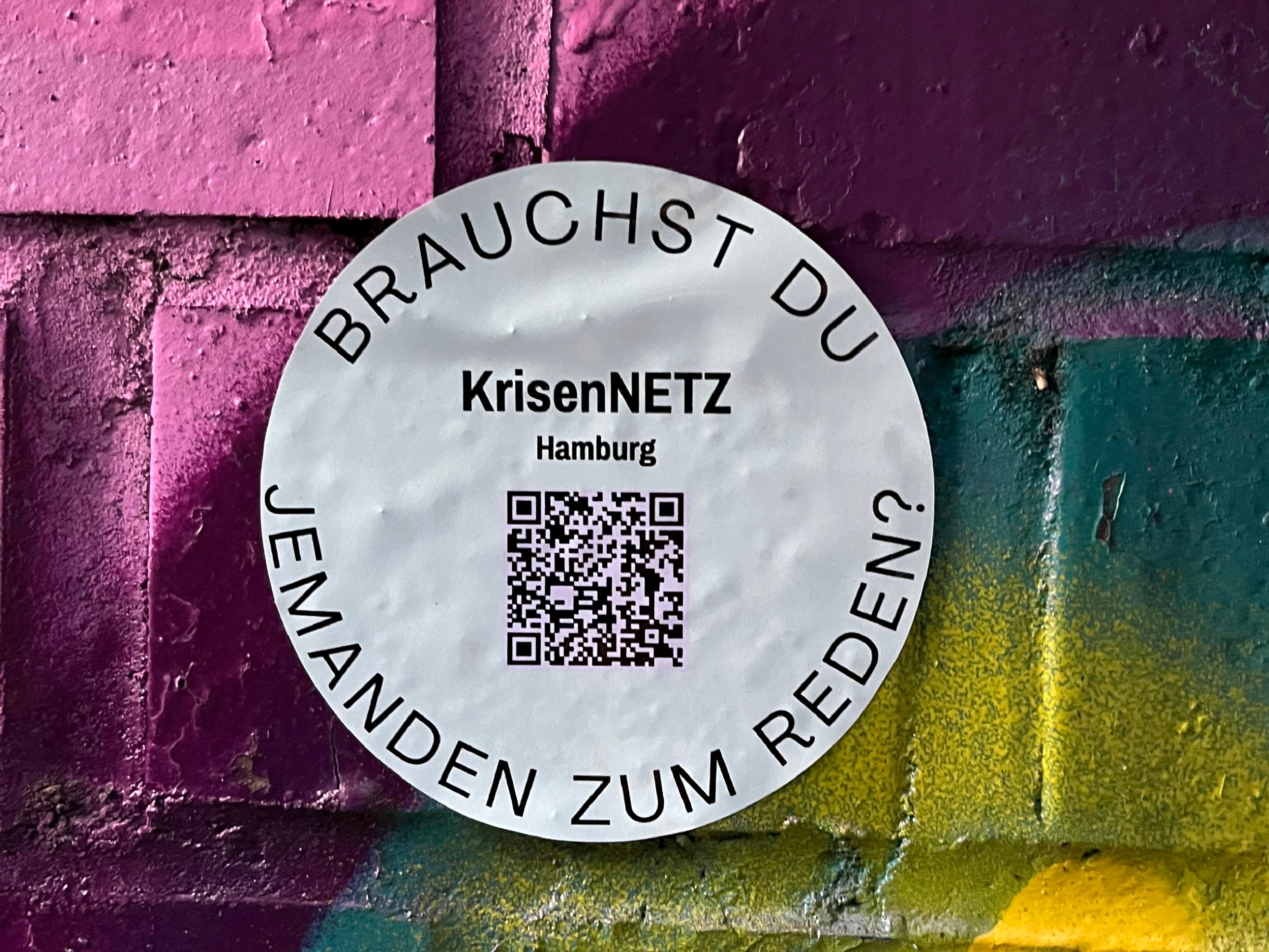 Runder Sticker mit QR-Code klebt an einer bunten, bemalten Ziegelwand; darauf steht KrisenNETZ Hamburg.