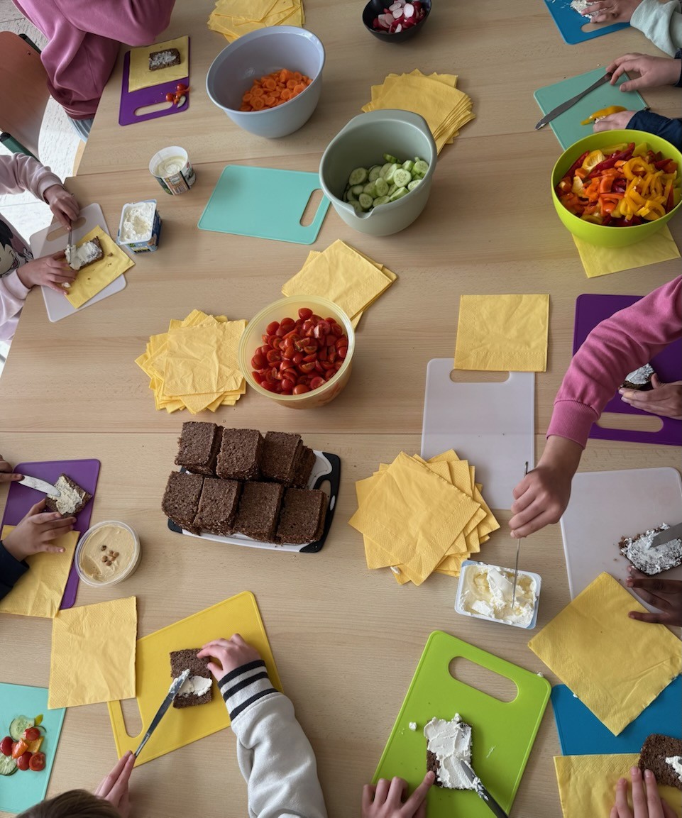 Mehrere Kinder bereiten belegte Brote zu, mit Käsecreme, Brot und buntem Gemüse wie Gurken, Tomaten und Paprika.