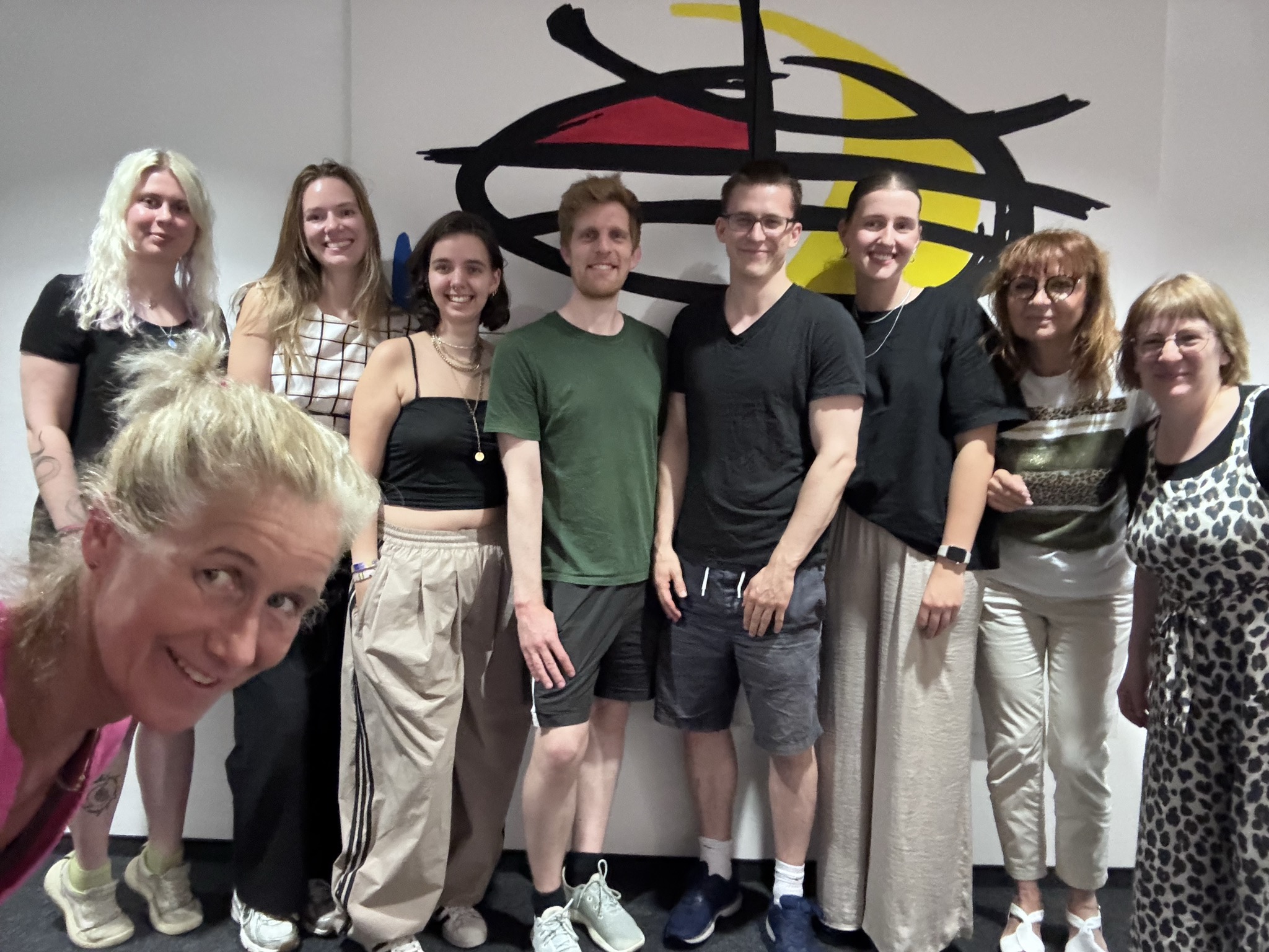 Gruppe von neun Menschen lächelt vor einer abstrakten Wandzeichnung mit schwarzen Linien und gelber Kreisscheibe.