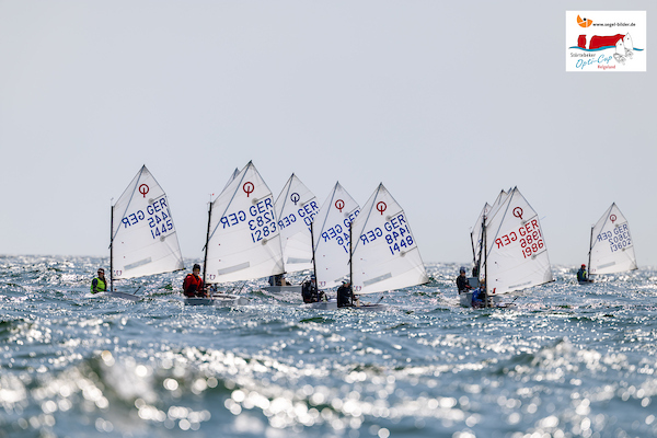 Mehrere kleine Jollen segeln bei einer Regatta dicht gedrängt auf dem Meer, Wellen glitzern im Sonnenlicht.