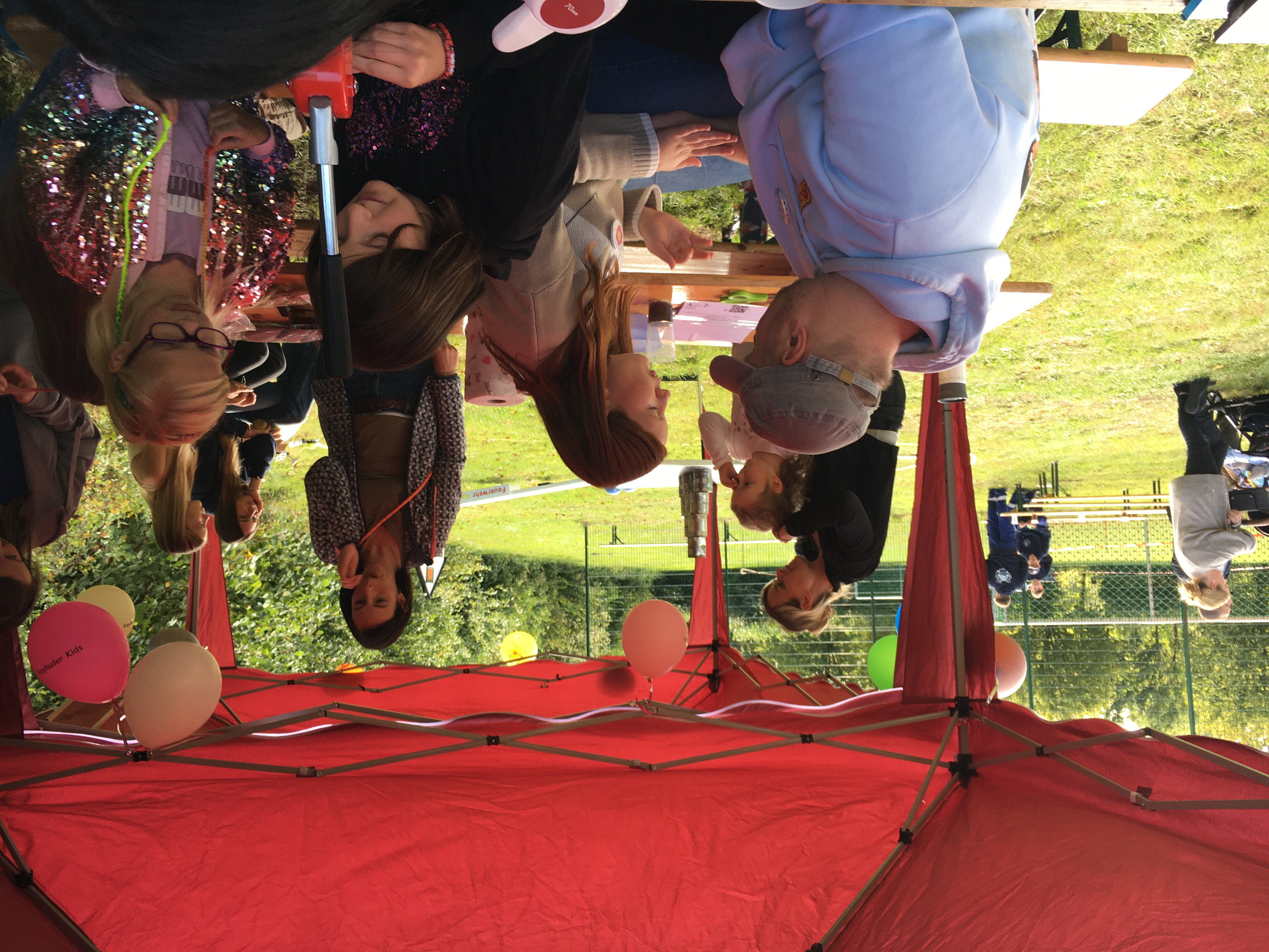 Menschen sitzen unter rotem Pavillon im Freien, Ballons hängen, Kinder basteln an Tischen.