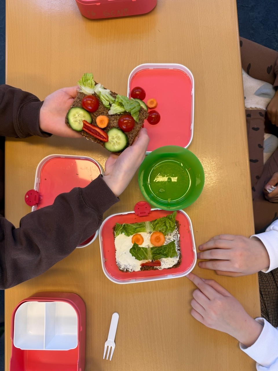 Zwei rote Lunchboxen mit Gemüse-Gesichtern aus Gurke, Tomate und Karotte auf einem Holztisch.