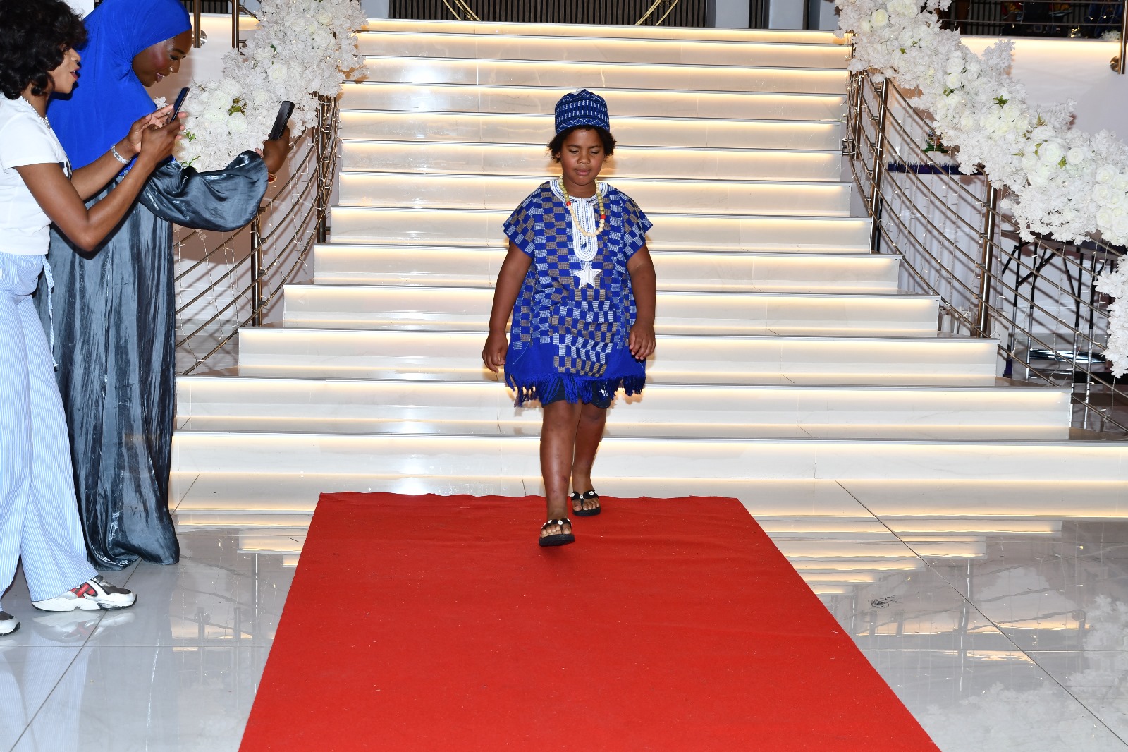 Ein Kind in blau kariertem Kleid mit Kopfbedeckung geht den roten Teppich hinunter, flankiert von Zuschauern und weißen Blumen.