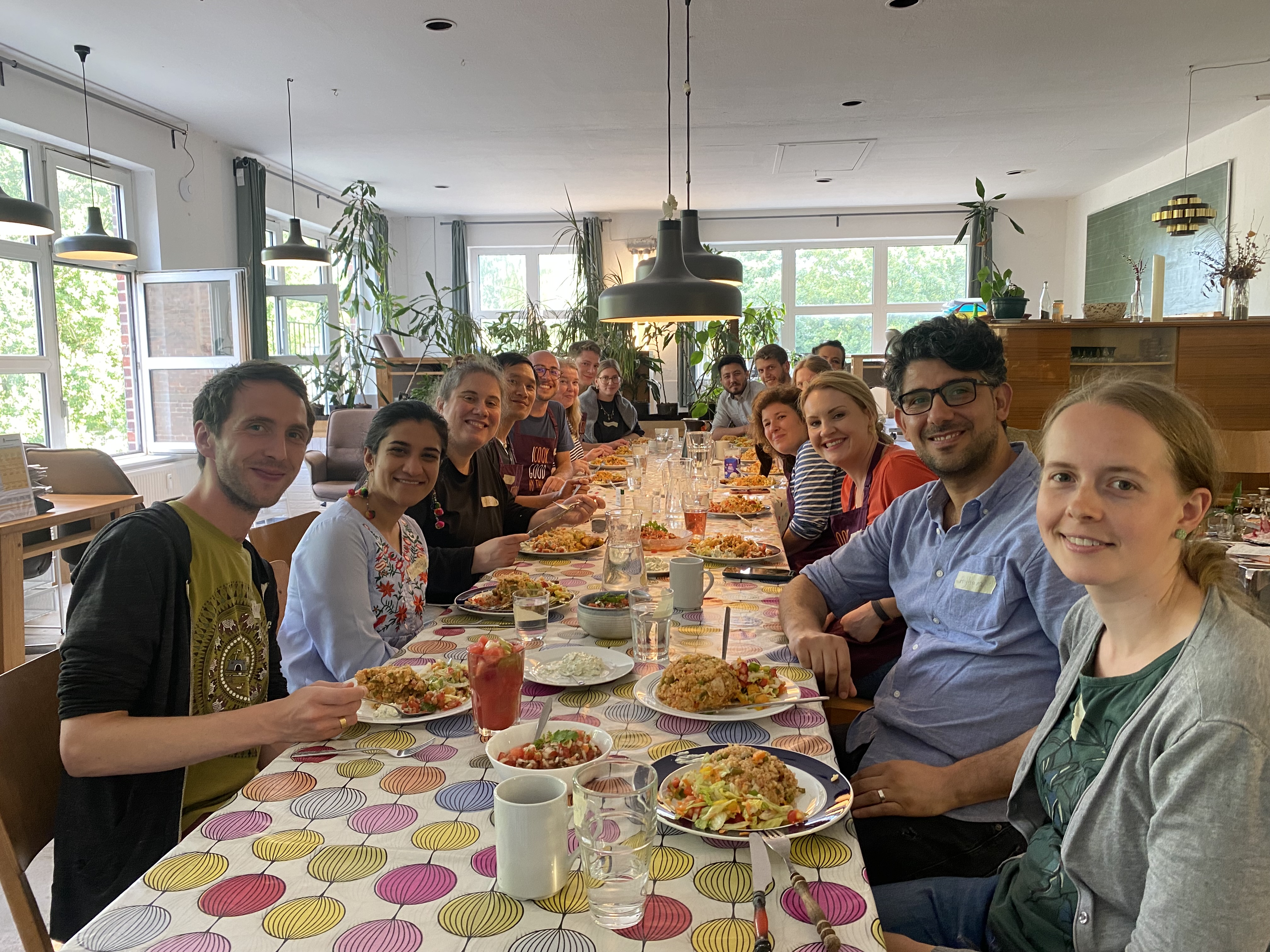 Eine Gruppe Menschen sitzt an einem langen Esstisch, isst, lacht und genießt Essen in einem hellen Raum voller Pflanzen.