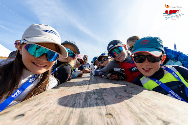 Gruppe Kinder und Jugendliche lehnt sich über einen Holztisch im Freien, alle tragen Sonnenbrillen und Caps.