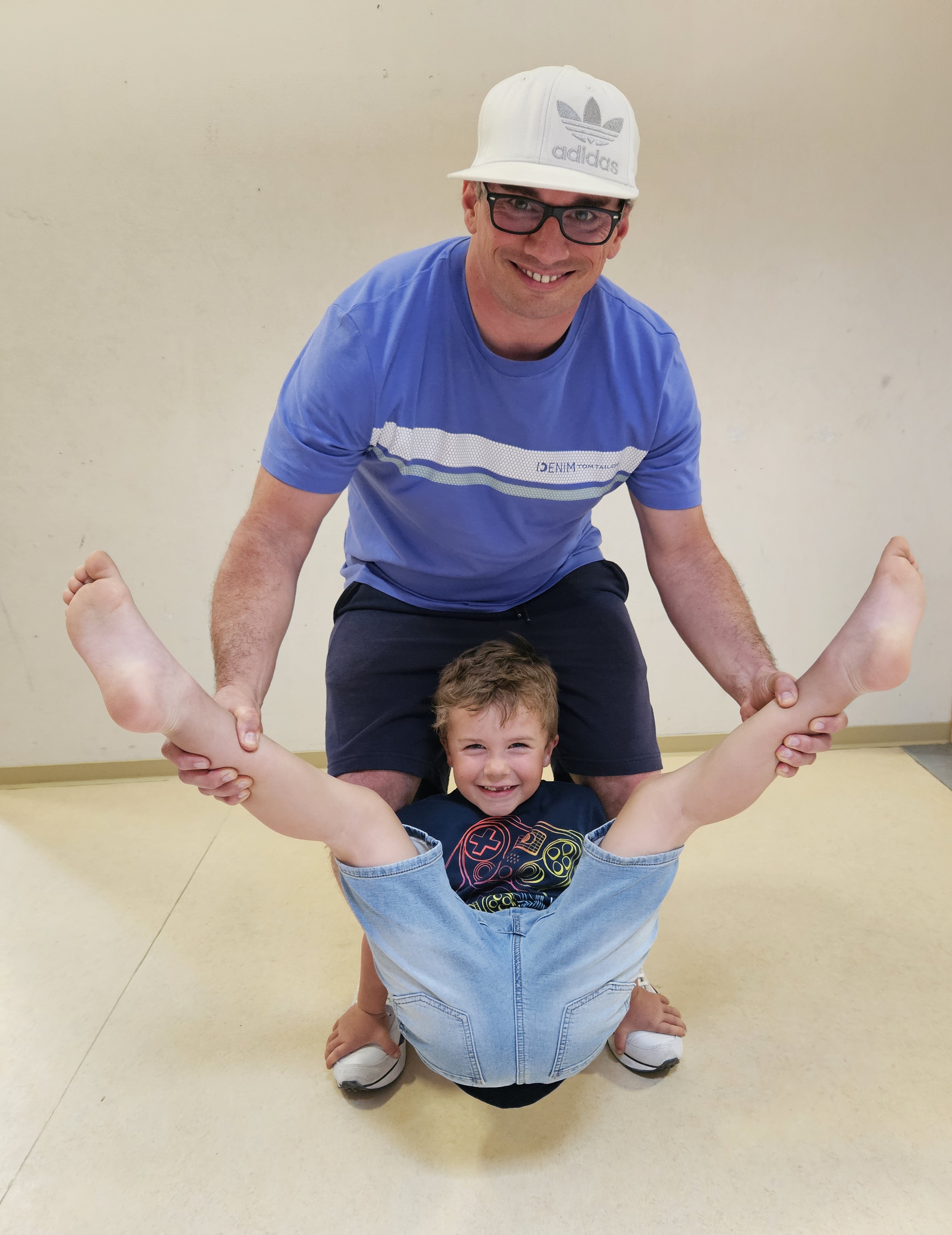 Mann mit weißem Adidas-Cap und Brille trägt blaues Shirt; lachender Junge hängt kopfüber zwischen seinen Beinen.