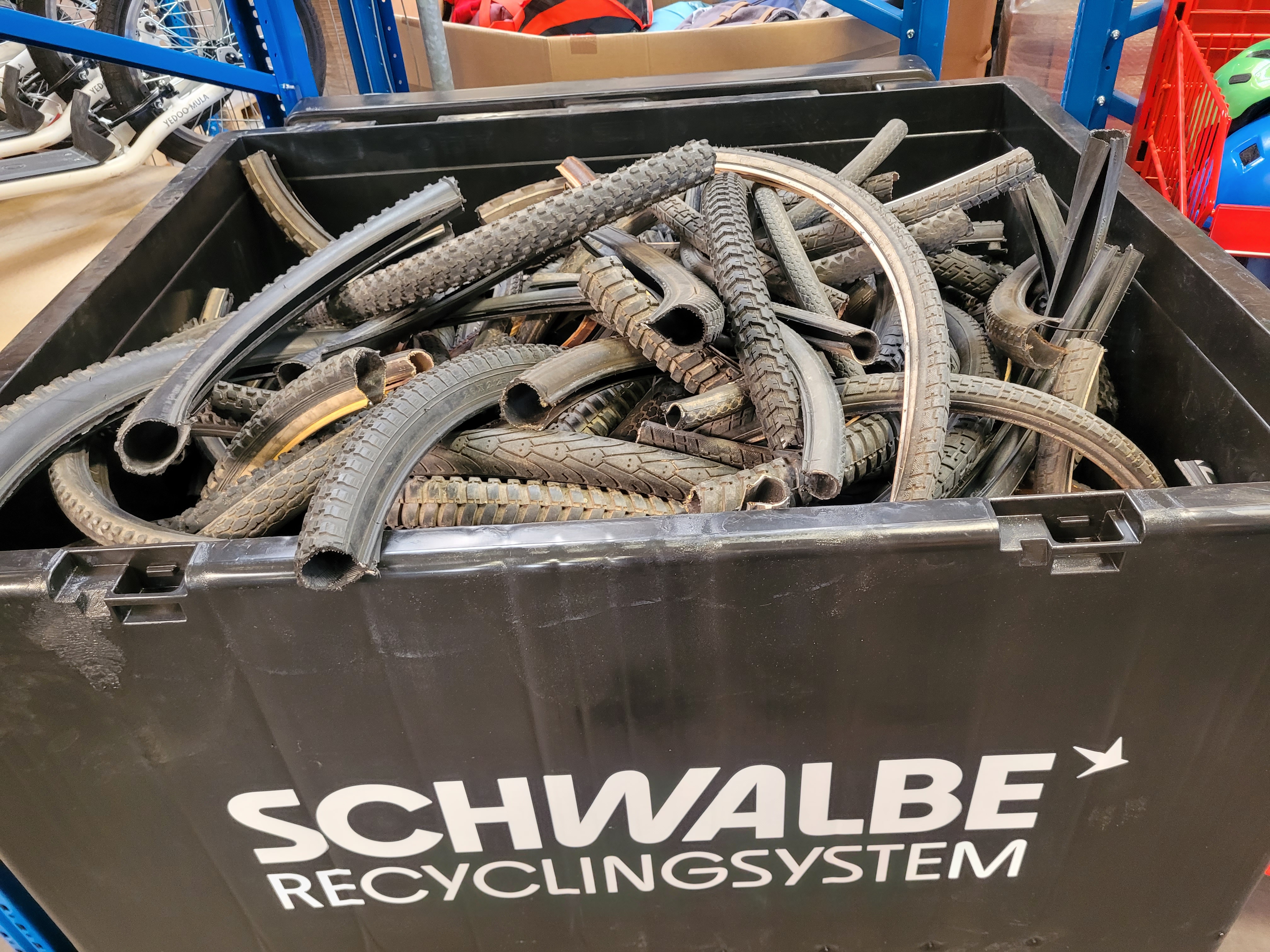 Großer schwarzer Behälter voller alter Fahrradreifen und Schläuche, Hintergrund mit Fahrrädern und blauem Regalsystem.