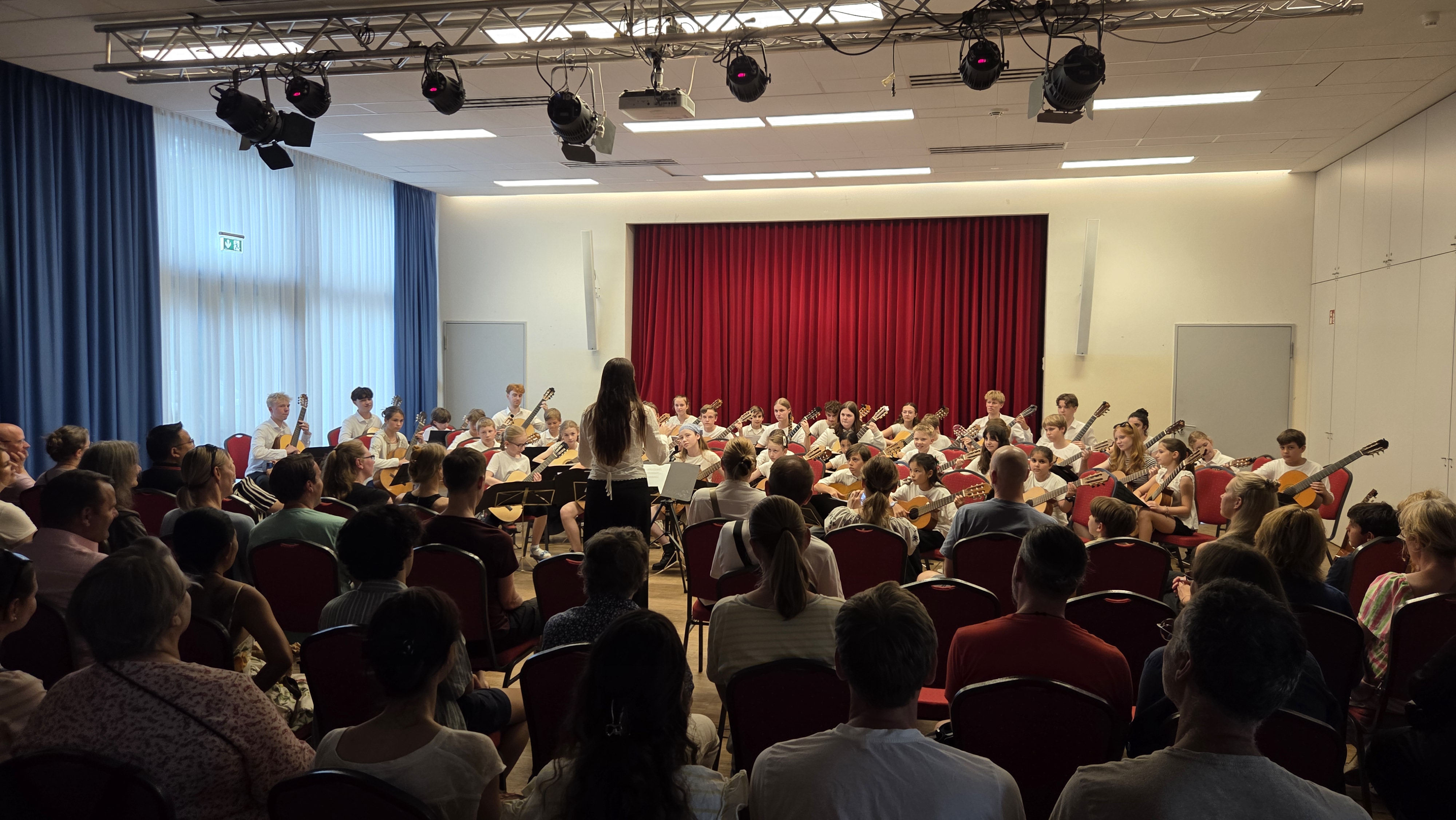 Viele Kinder mit Gitarren sitzen auf der Bühne, Dirigentin führt, roter Vorhang im Hintergrund, Publikum schaut.