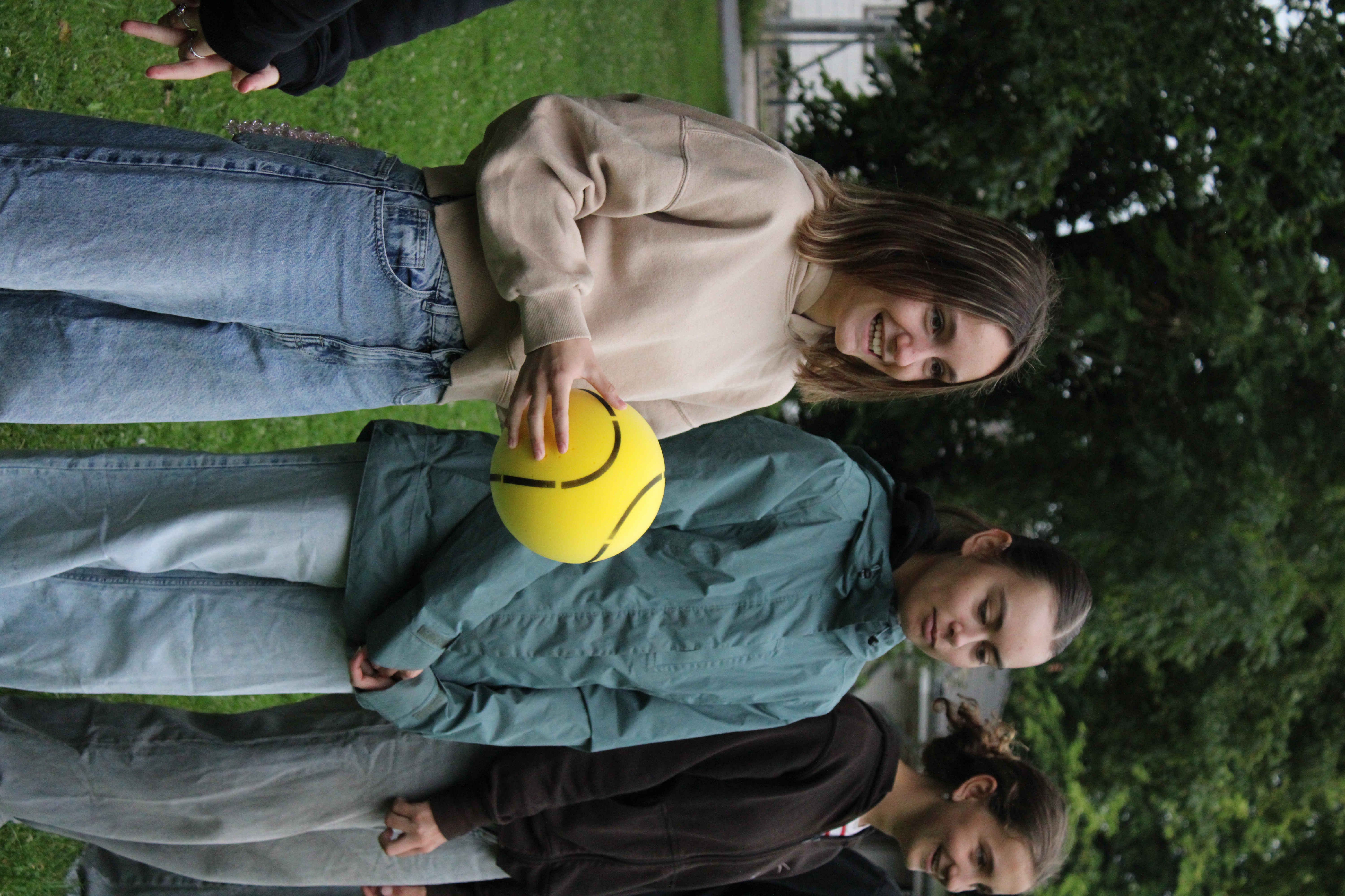 Drei Mädchen stehen zusammen im Park; die vorderste hält einen gelben Ball und lächelt.