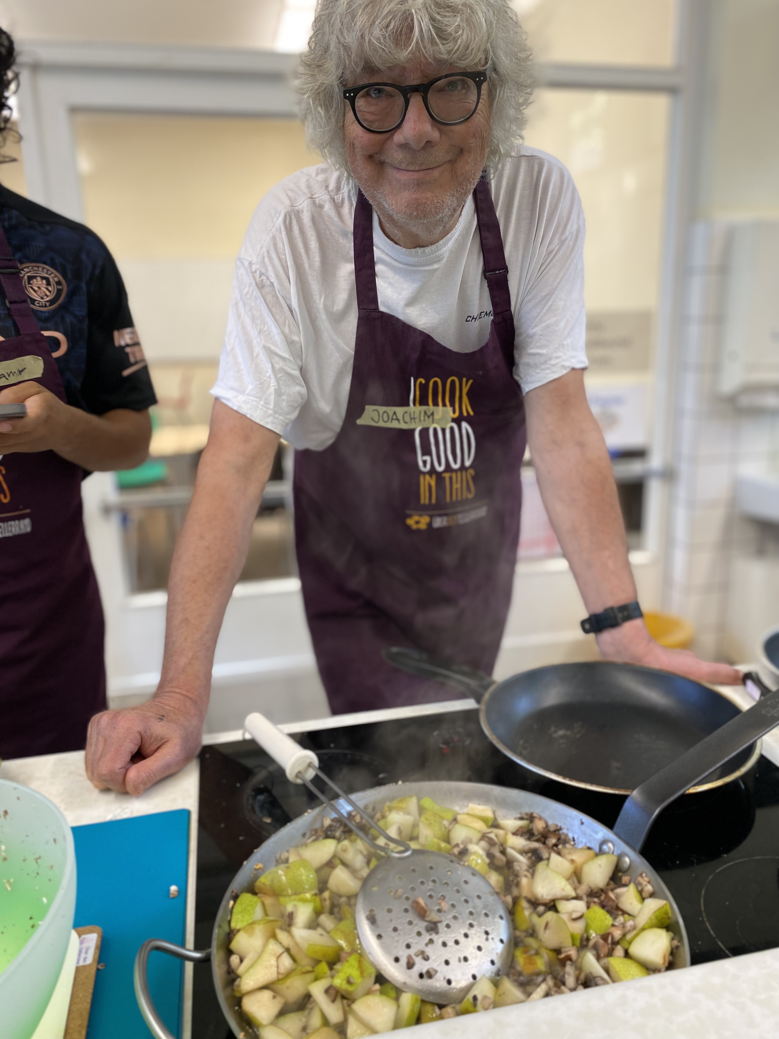 Älterer Mann mit grauen Haaren und Brille in der Küche, Schürze an, rührt in Pfanne mit gewürfeltem Obst.