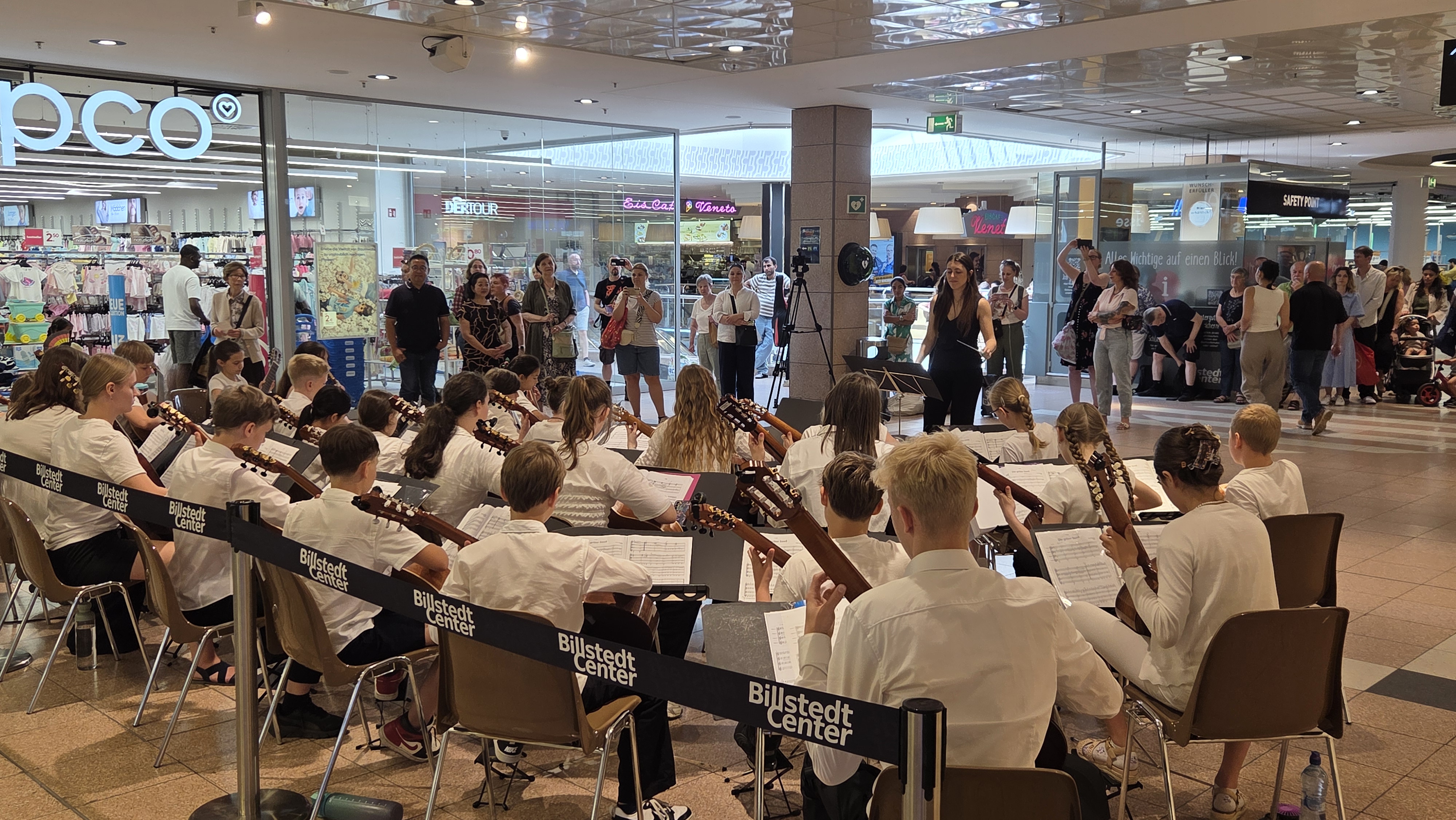 Jugendorchester mit Gitarren spielt im Einkaufszentrum vor Publikum; Geschäfte und Glasfronten im Hintergrund.