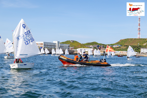 Viele kleine Segelboote auf dem Wasser, ein rotes Motorboot mit zwei Personen fährt vorbei, Küste mit Hügeln im Hintergrund.
