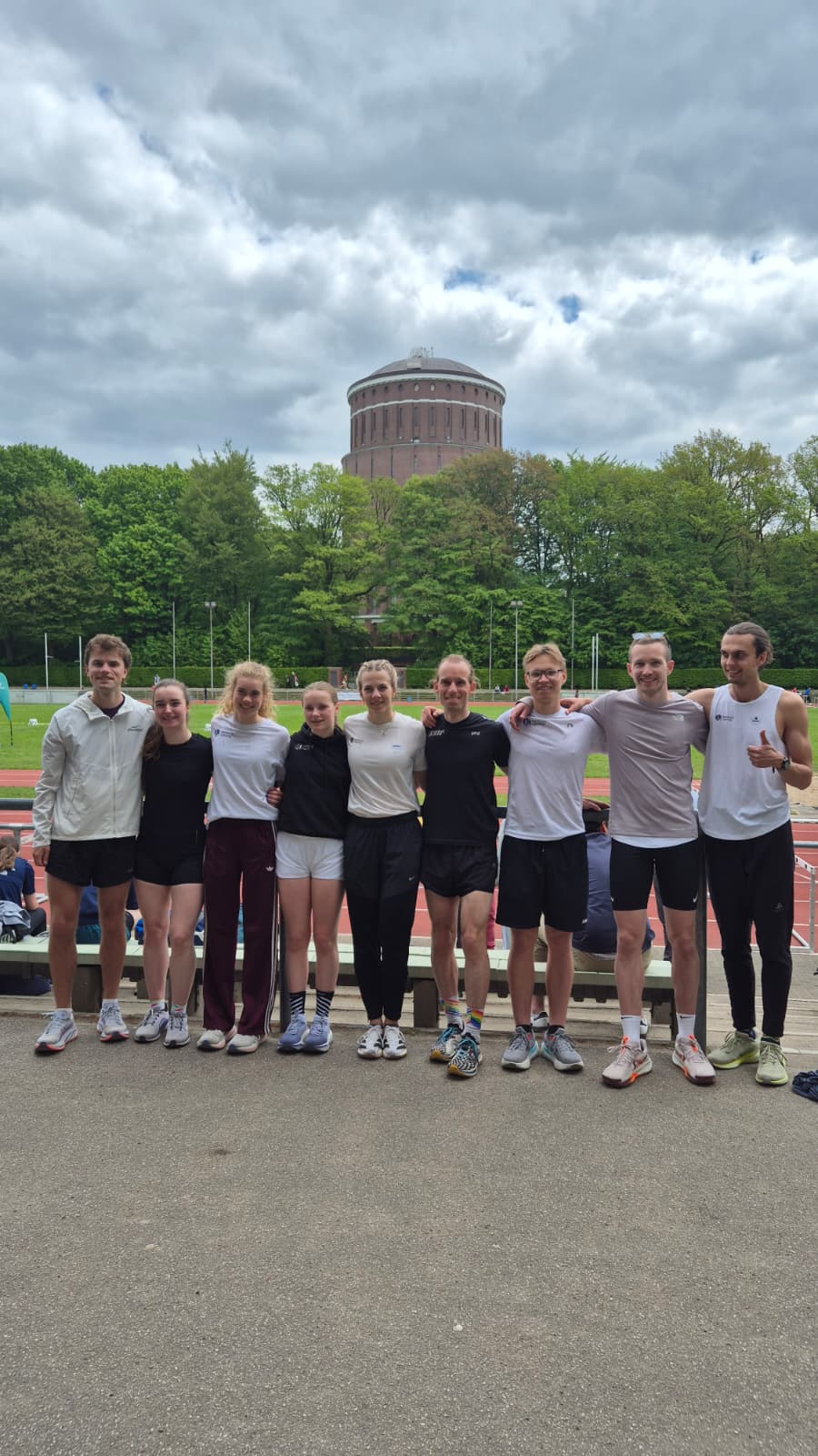 Gruppe junger Athleten steht am Rand einer Laufbahn vor einem runden Backsteinturm und grünen Bäumen.