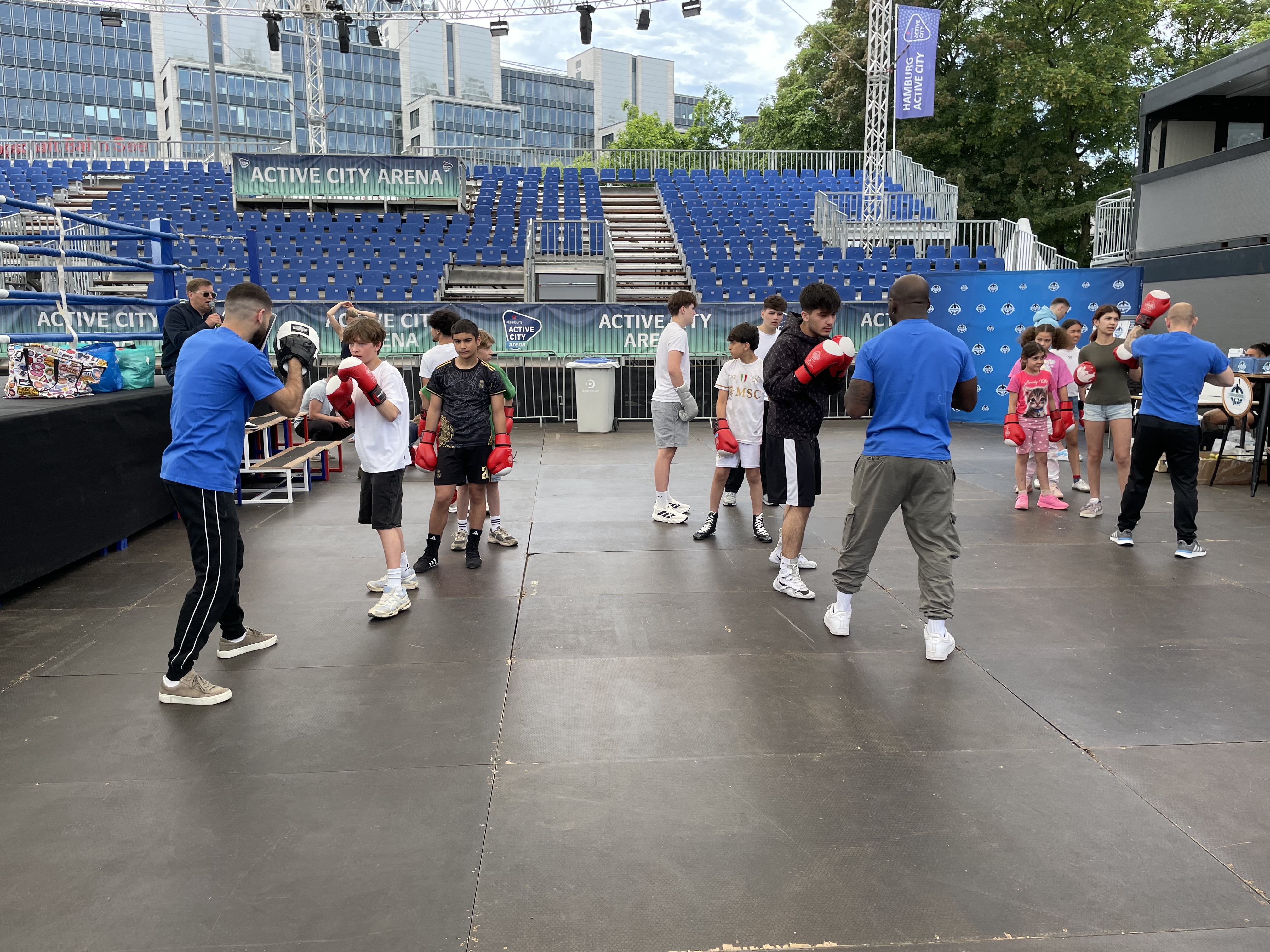 Kinder boxen mit Trainern in der Active City Arena, blaue Tribünen und moderne Bürogebäude im Hintergrund.