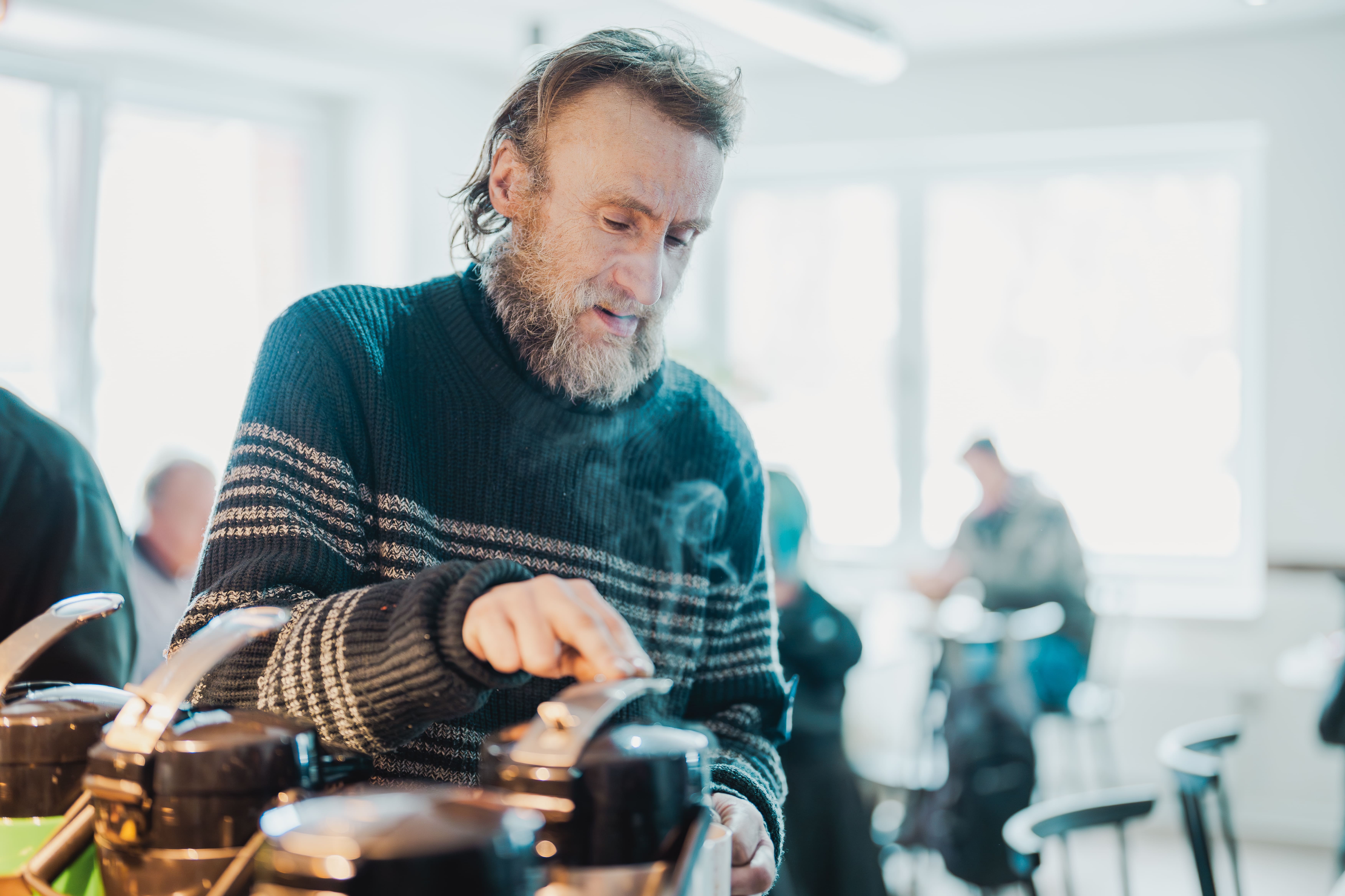 Ein älterer Mann mit Bart und gestricktem Pullover bearbeitet Metallkannen auf einem Tisch in einer hellen, modernen Küche.