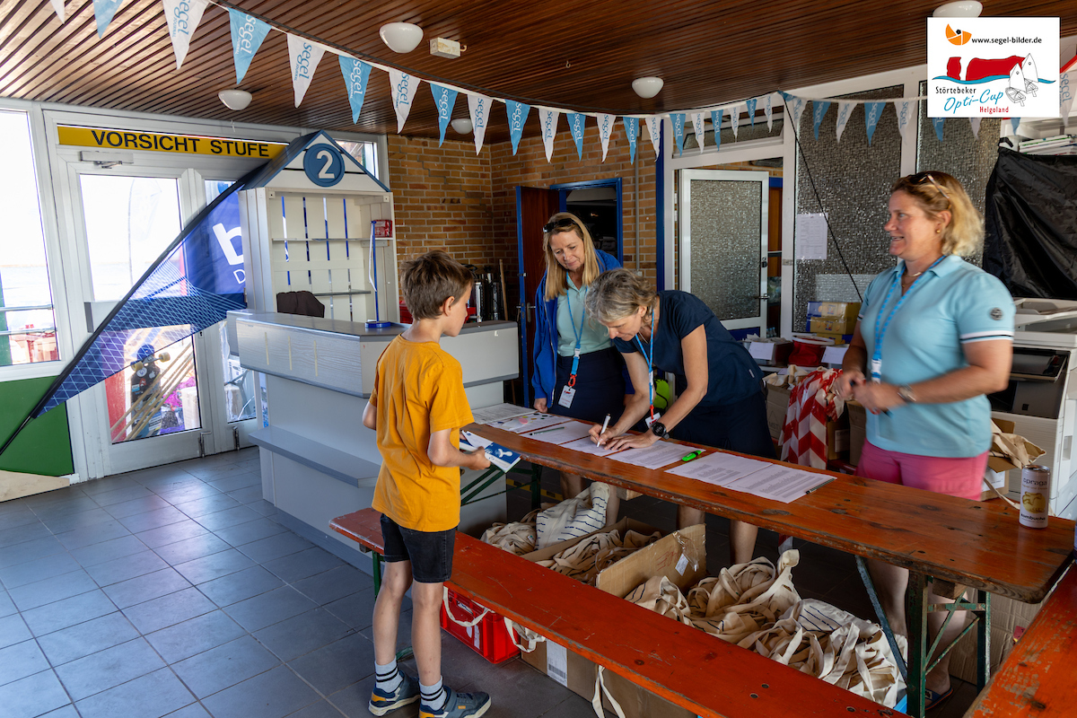 Ein Junge im orangefarbenen T‑Shirt steht am Check-in-Tisch, während drei Frauen Formulare ausfüllen.