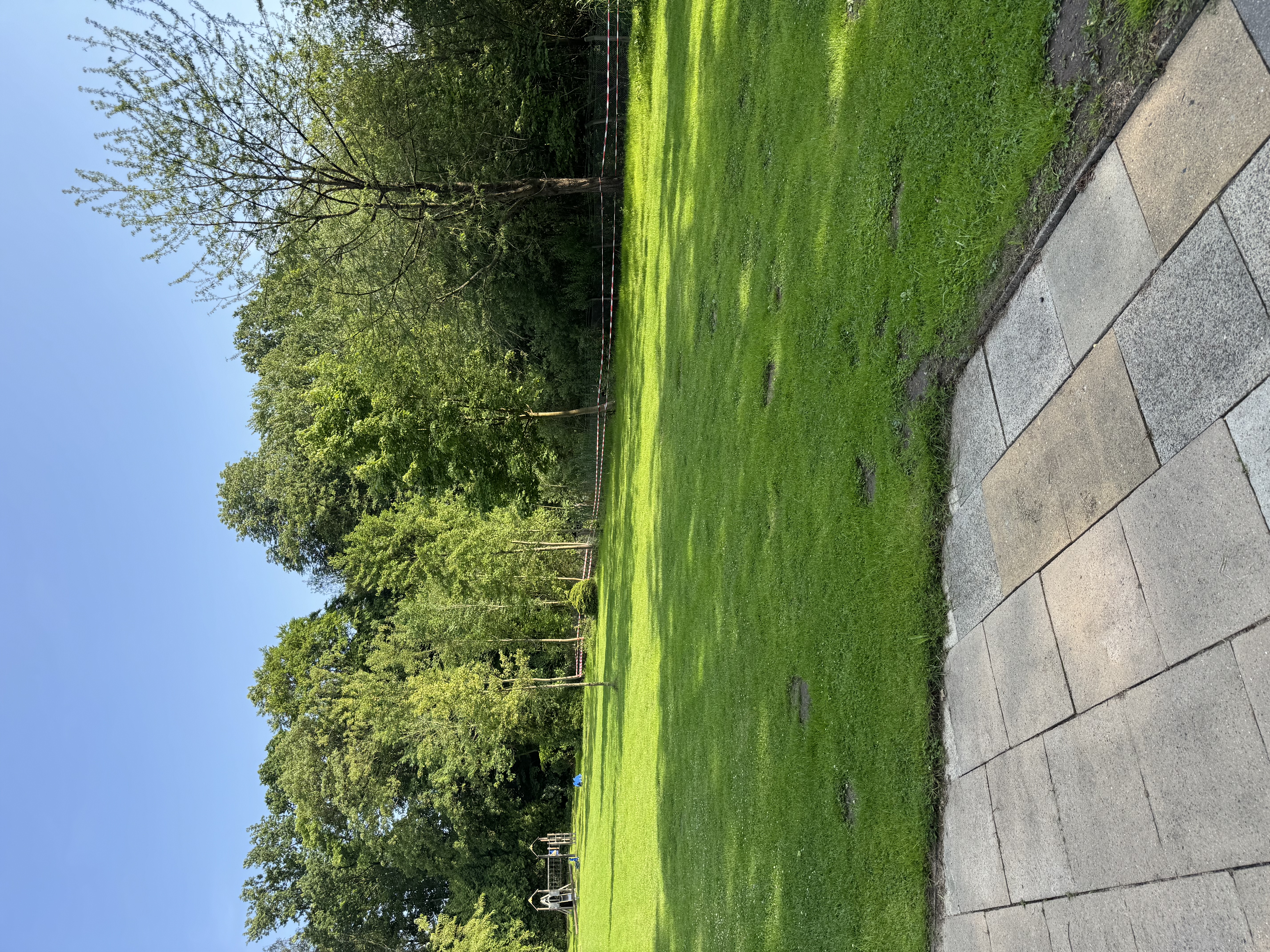 Grüne Parkfläche mit Spielgerät im Hintergrund, blauer Himmel, Vordergrund aus gepflasterten Steinplatten und Absperrband am