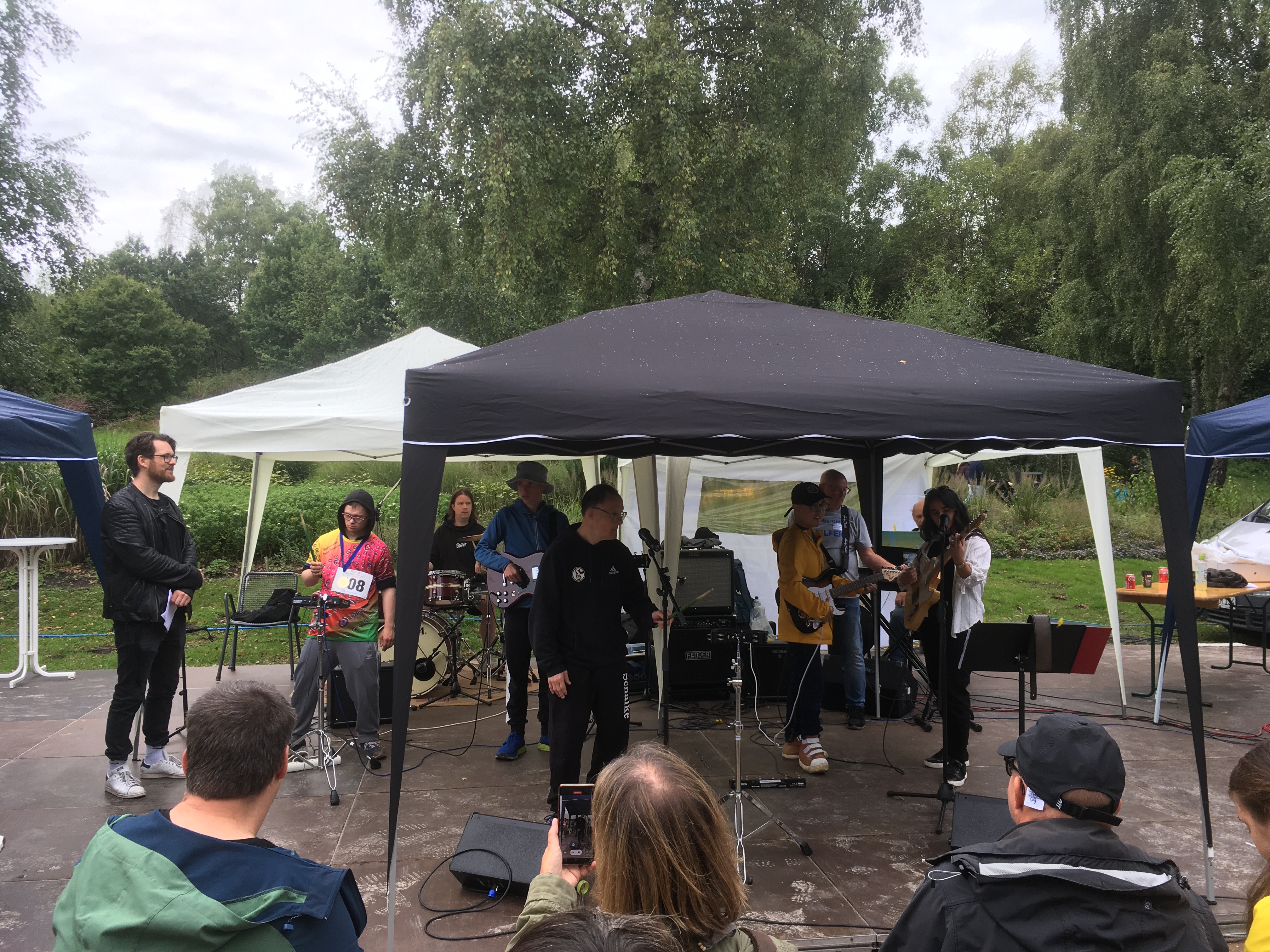 Musiker spielen draußen unter Zelten mit Gitarren, Schlagzeug und Mikrofonen, Zuschauer schauen zu.