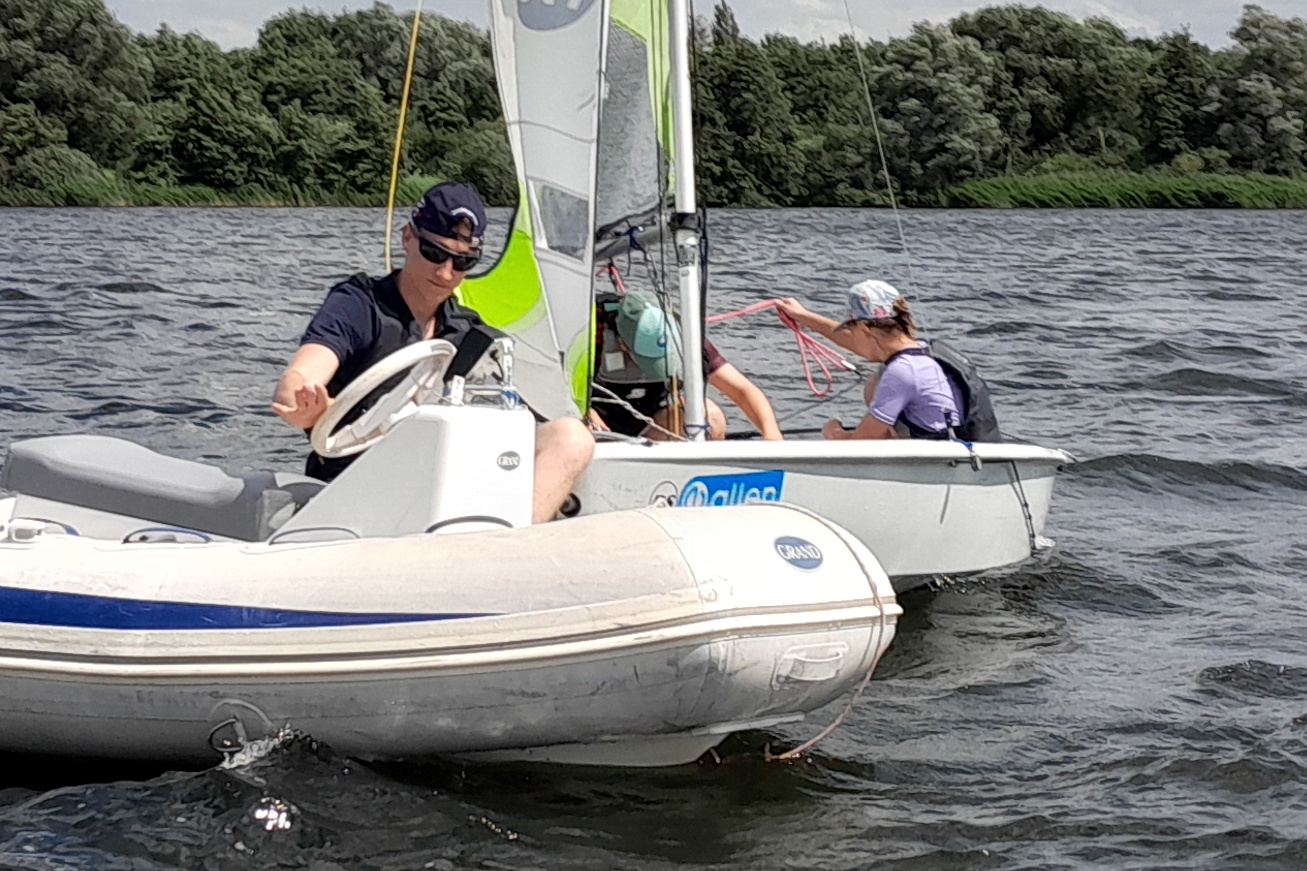 Zwei Segler auf einem kleinen Boot, einer steuert, die andere hält ein rotes Seil; Wasser und Ufer mit Bäumen dahinter.