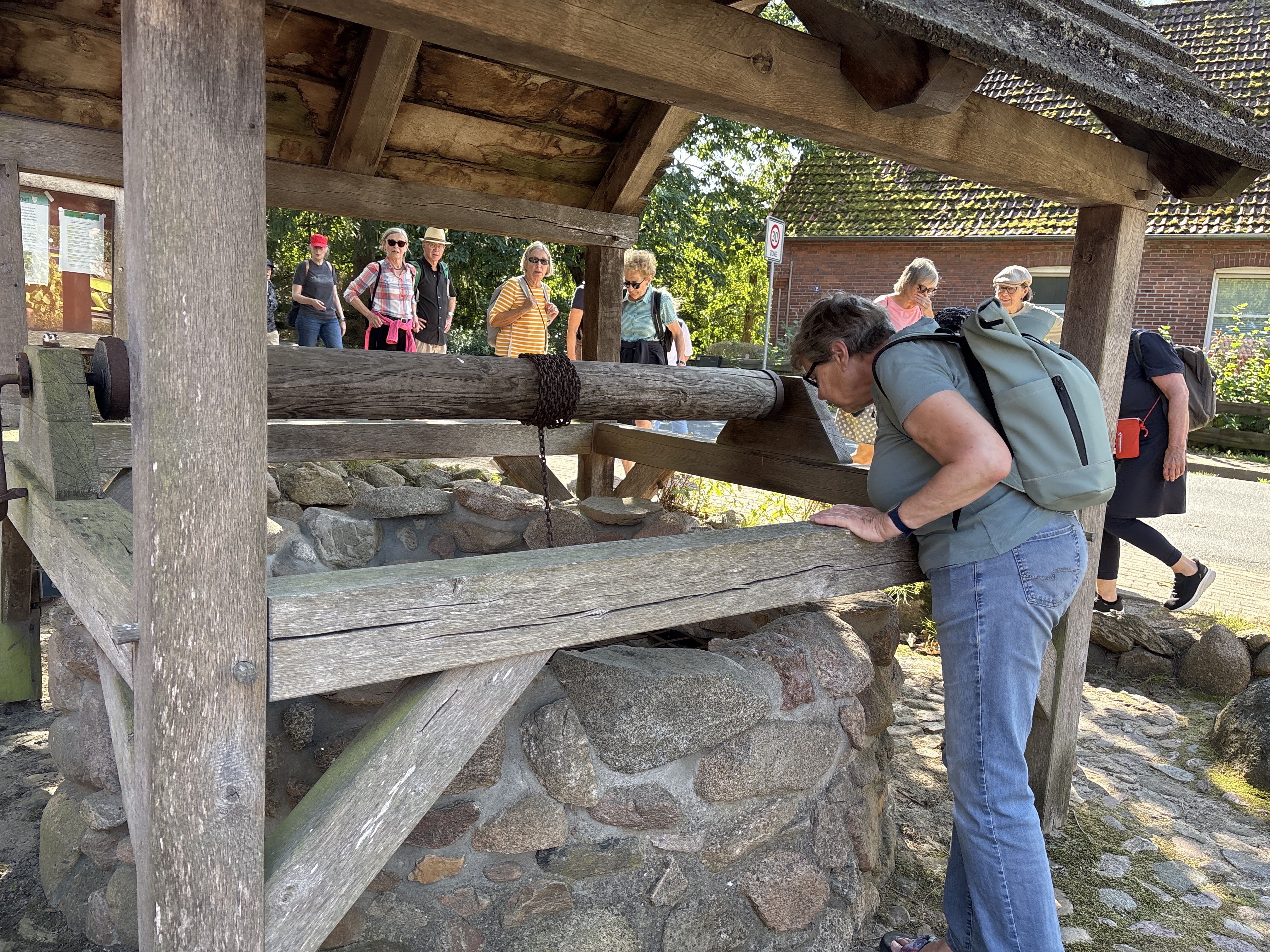Eine Gruppe älterer Menschen steht unter einem Holzdach und schaut auf einen Holztrog mit Steinfundament.