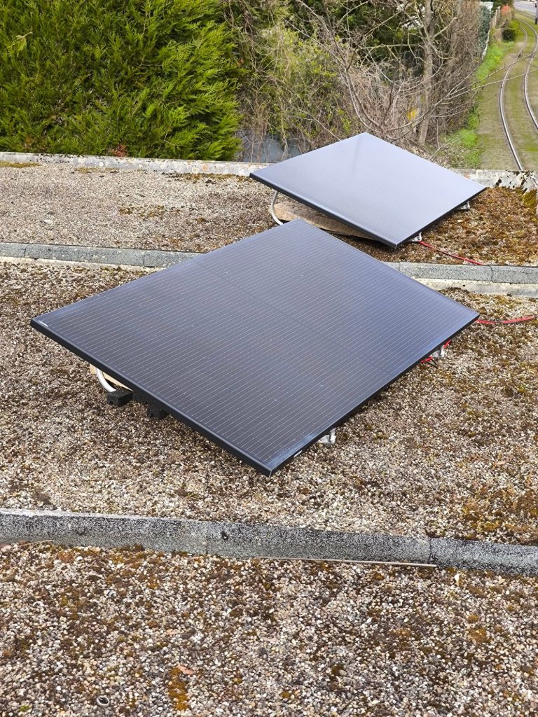 Zwei Solarpaneele liegen flach auf einem kiesbedeckten Flachdach, im Hintergrund sind Büsche und ein Gleisabschnitt zu sehen.