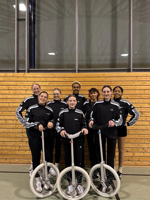 Gruppe von Sportlern in schwarzen Trainingsjacken posiert mit Einrädern vor einer Holzwand im Gym.