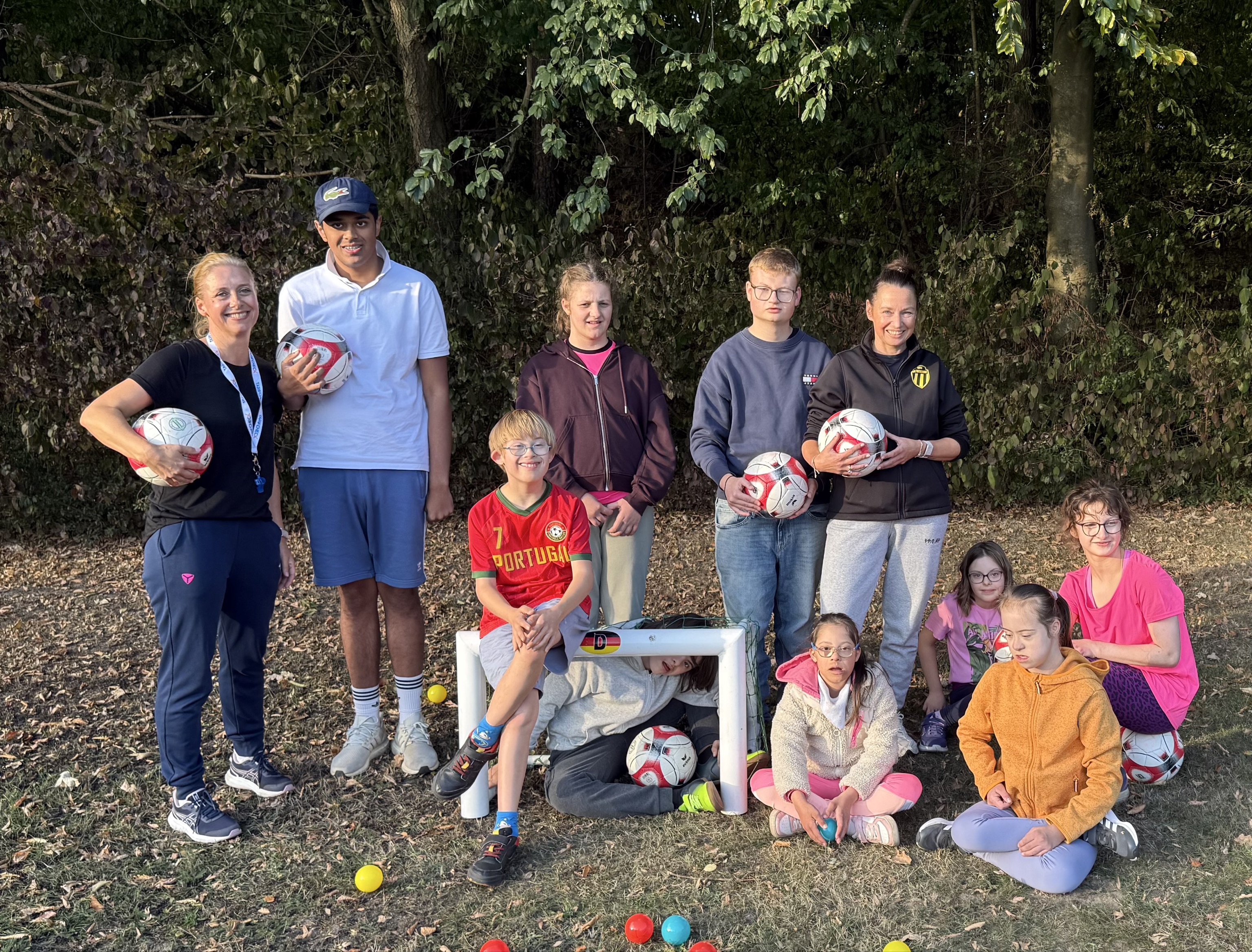 Gruppe Erwachsener und Kinder posiert draußen mit Fußbällen vor einer dichten Hecke.