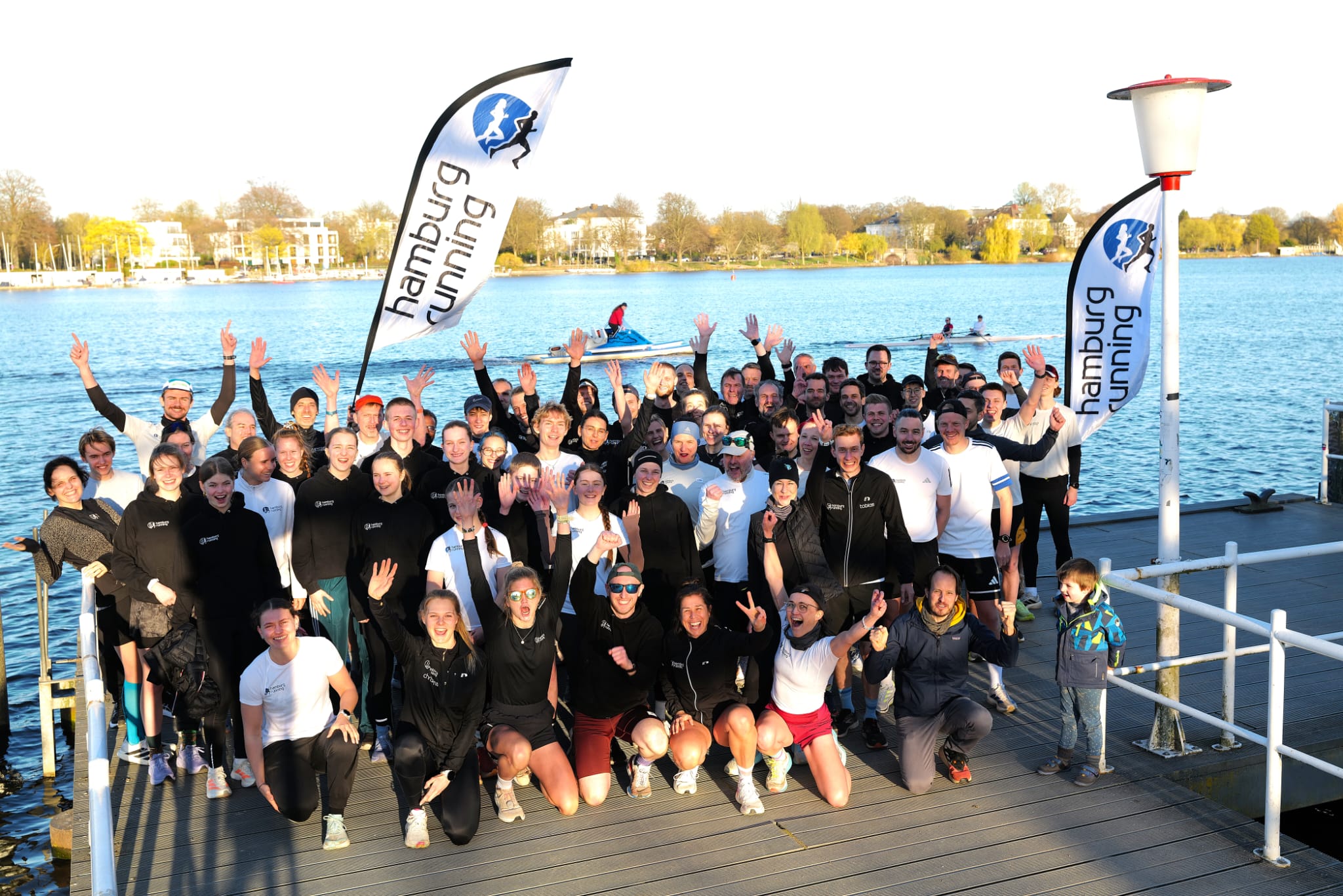 Große Gruppe Läuferinnen und Läufer winkt jubelnd vom Pier am See, Banner „Hamburg Running“ weht im Hintergrund.