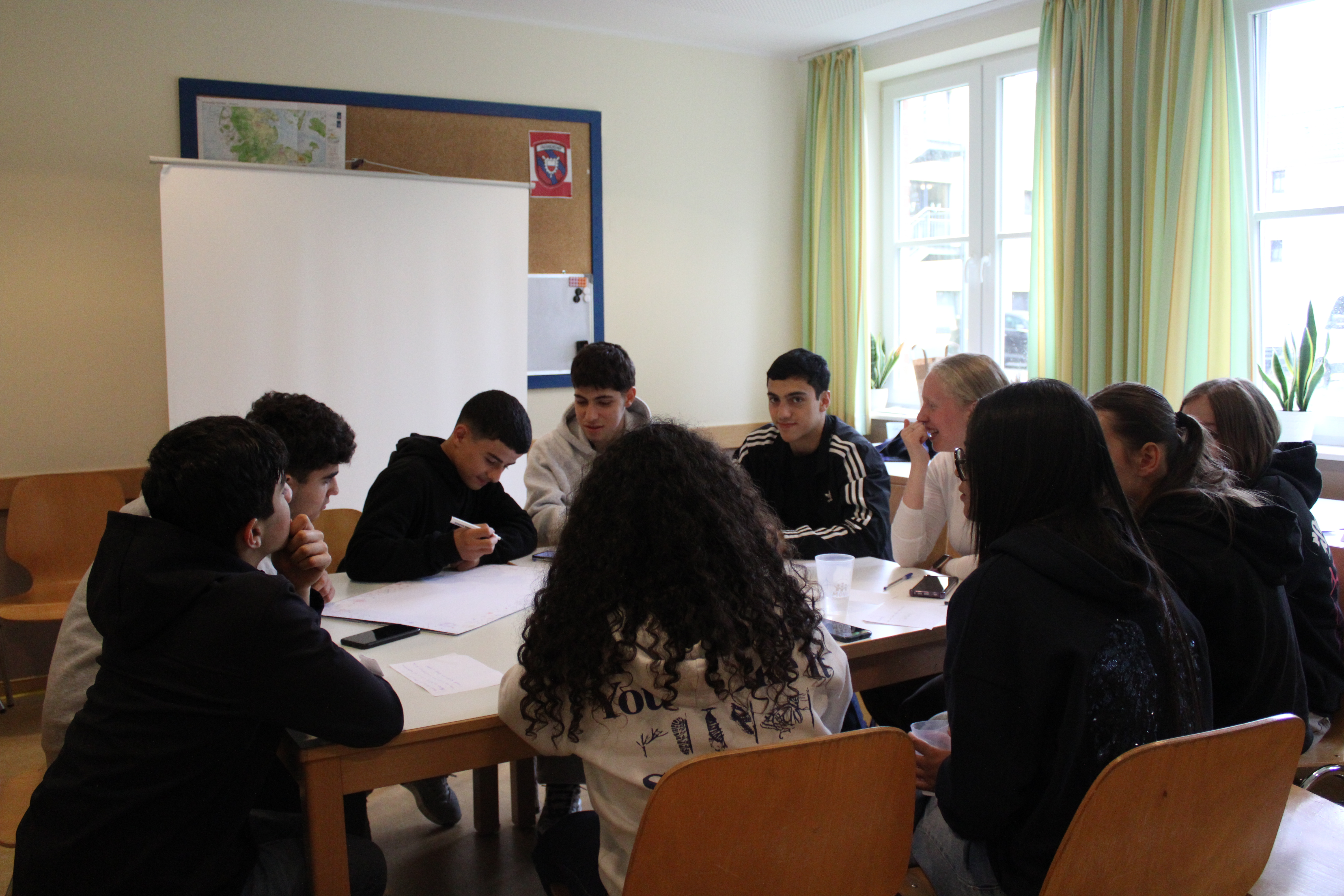 Gruppe von Schülern sitzt an Tischen in einem Klassenraum, diskutiert und schreibt, hinter ihnen Fenstern mit Vorhängen.