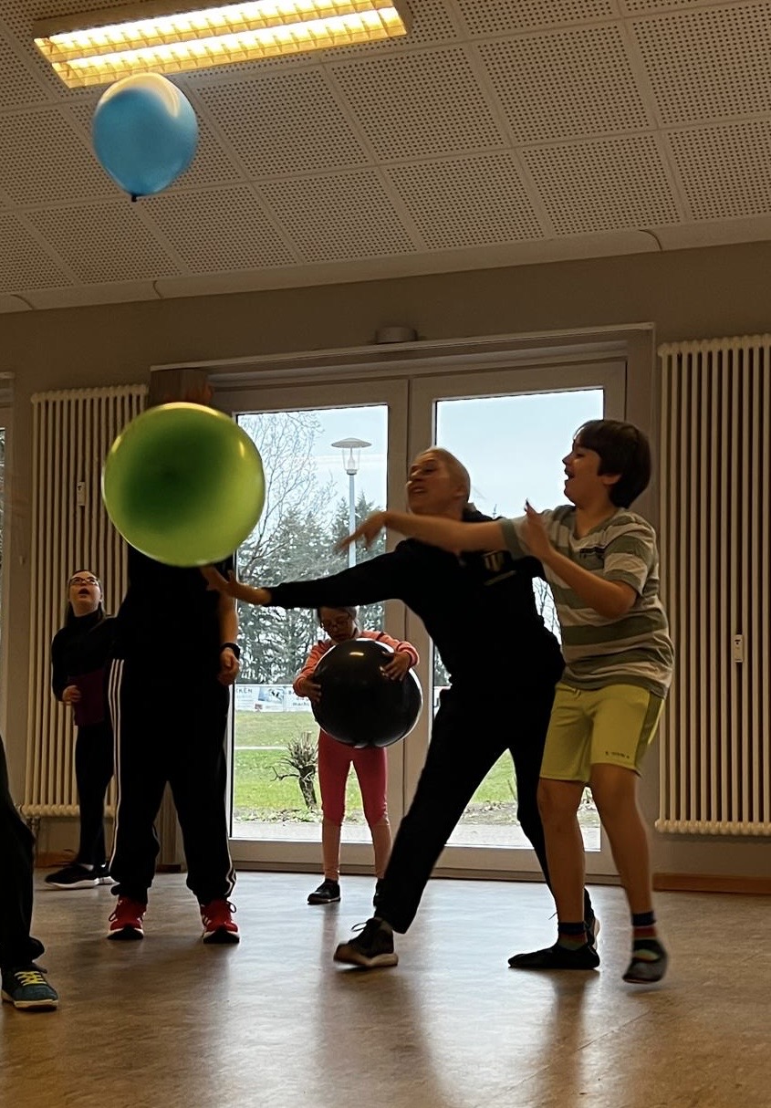 Mehrere Kinder spielen drinnen mit bunten Luftballons, vor großen Fenstern mit Blick nach draußen.