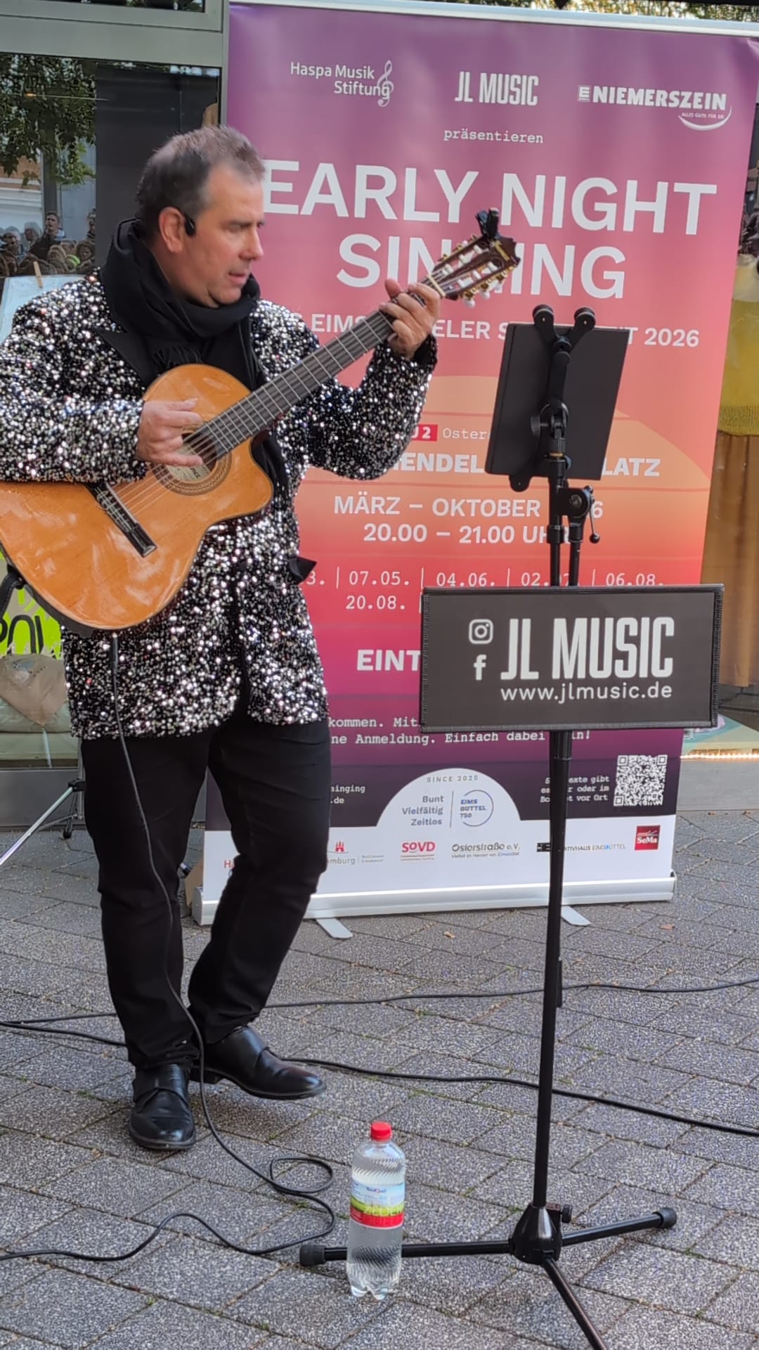 Ein Musiker mit Glitzerjacke spielt Gitarre vor einem bunten Banner mit der Aufschrift Early Night Singing.