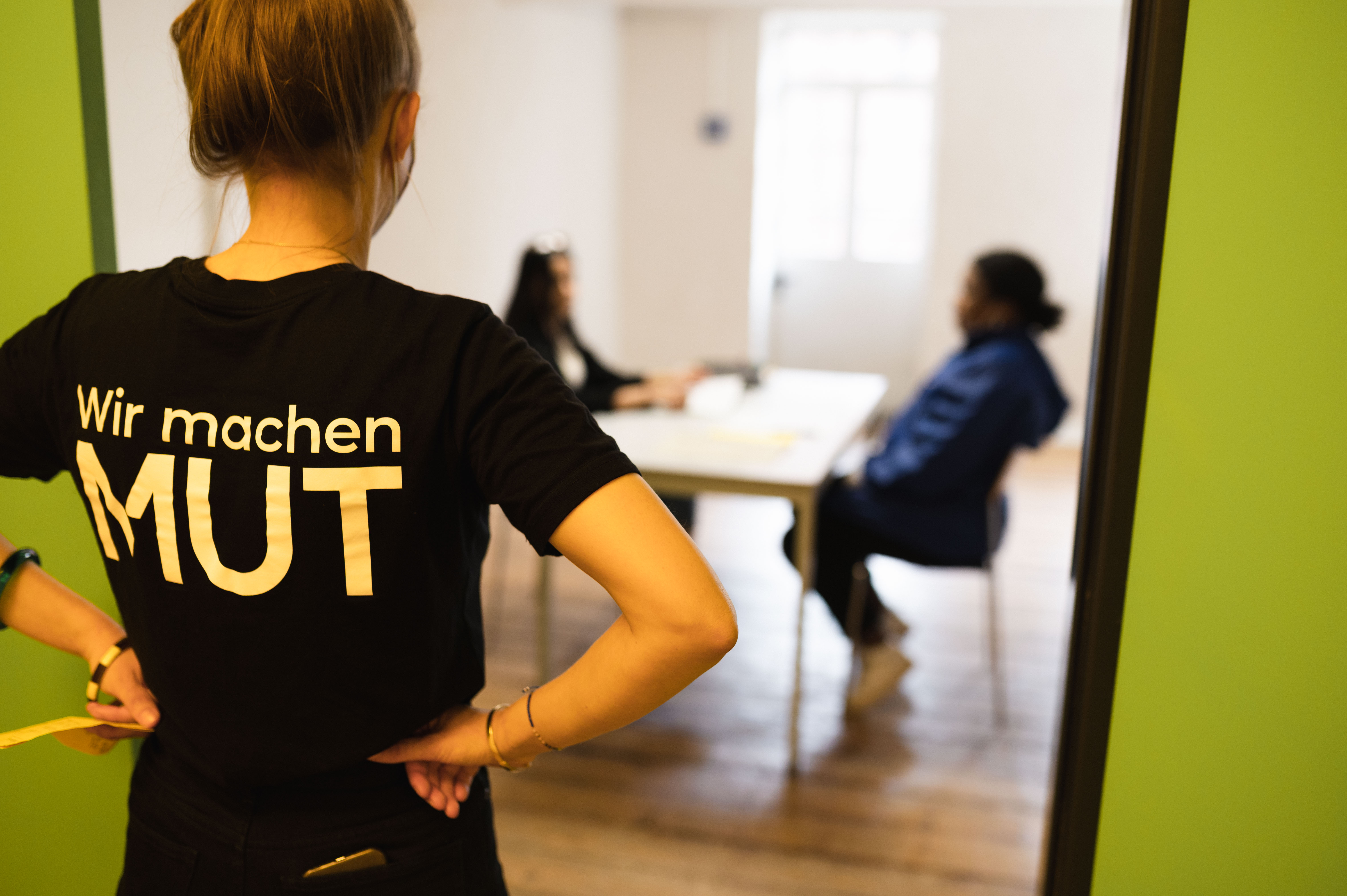 Person von hinten in schwarzem Shirt vor grünem Türrahmen, im Hintergrund zwei Personen am Tisch.