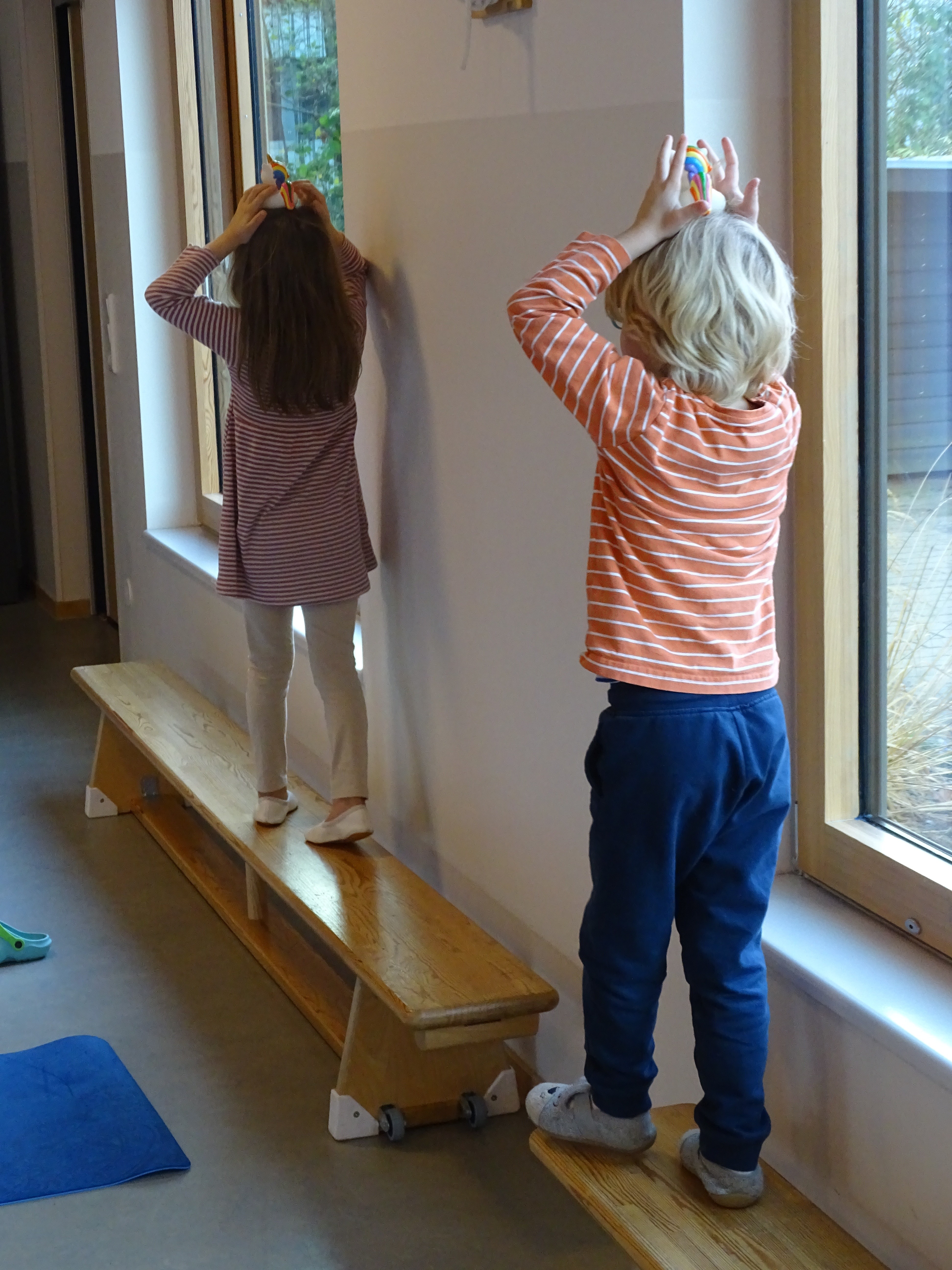 Zwei Kinder balancieren auf einer Holzstange und halten bunte Spielzeugfiguren über dem Kopf.