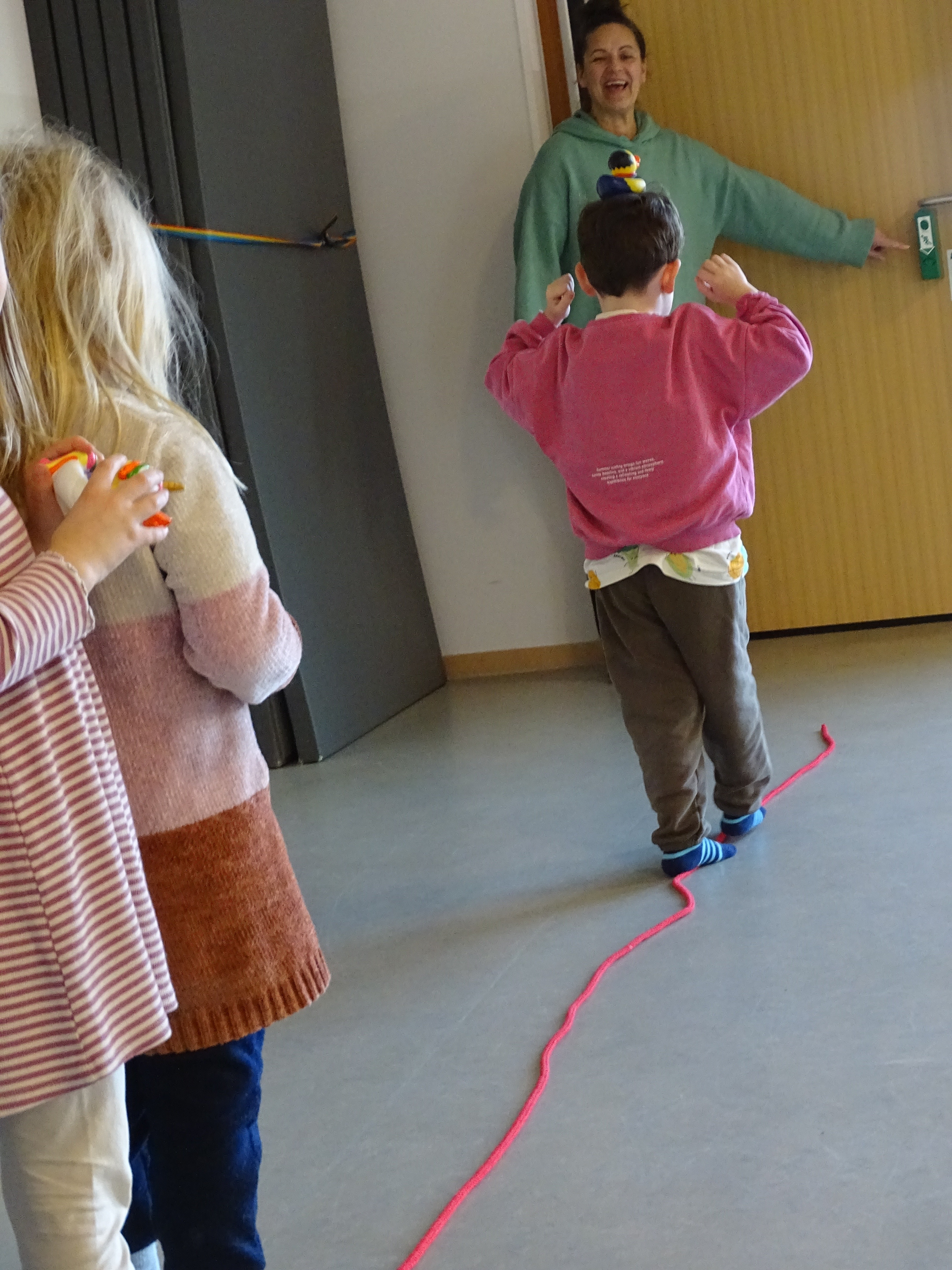 Eine Frau in grünem Hoodie lächelt, ein Kind in pinkem Sweatshirt von hinten, eine pinke Schnur liegt auf dem Boden.