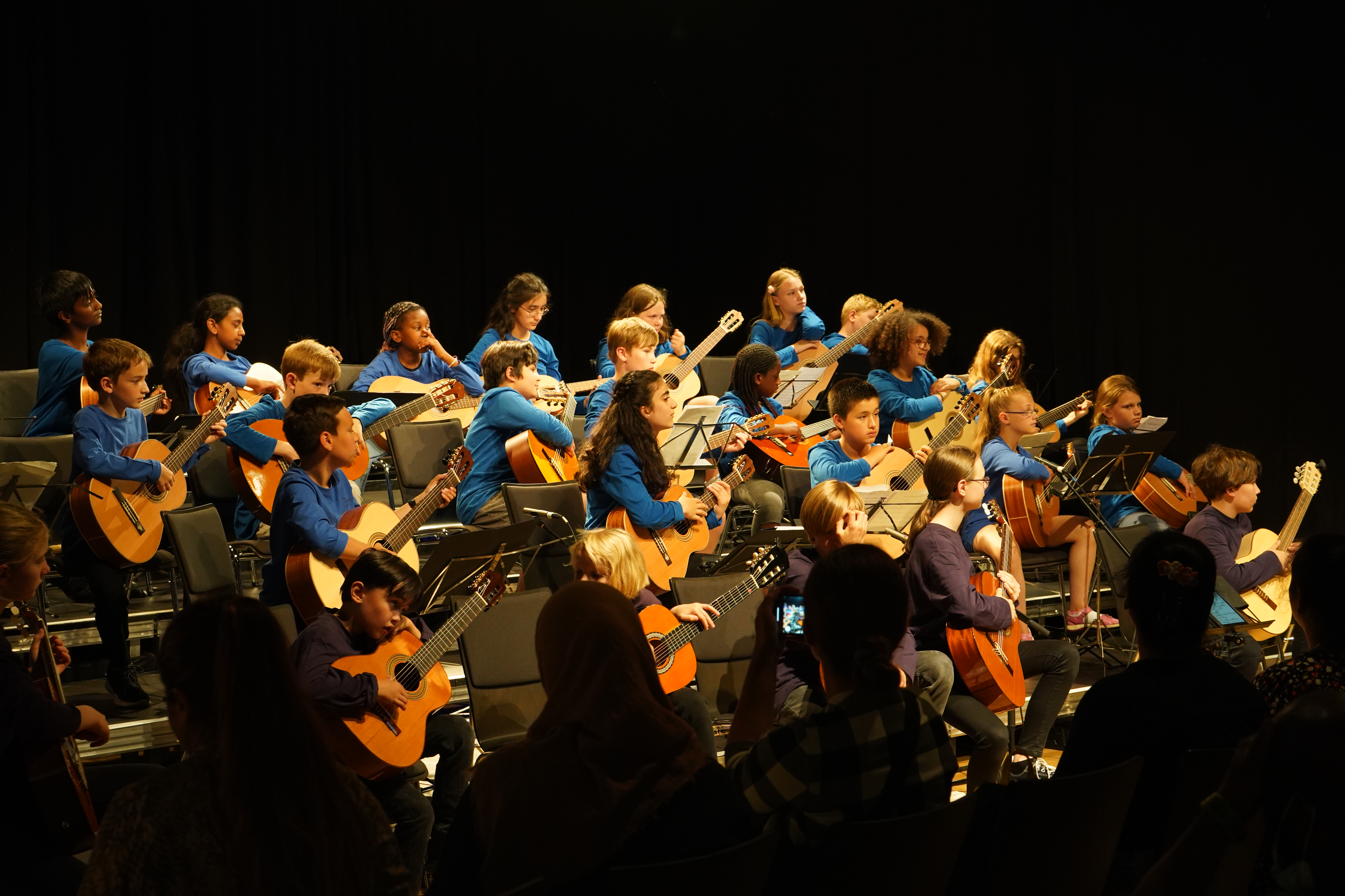 Viele Kinder in blauen Shirts spielen Gitarren in einem Orchester auf der Bühne.
