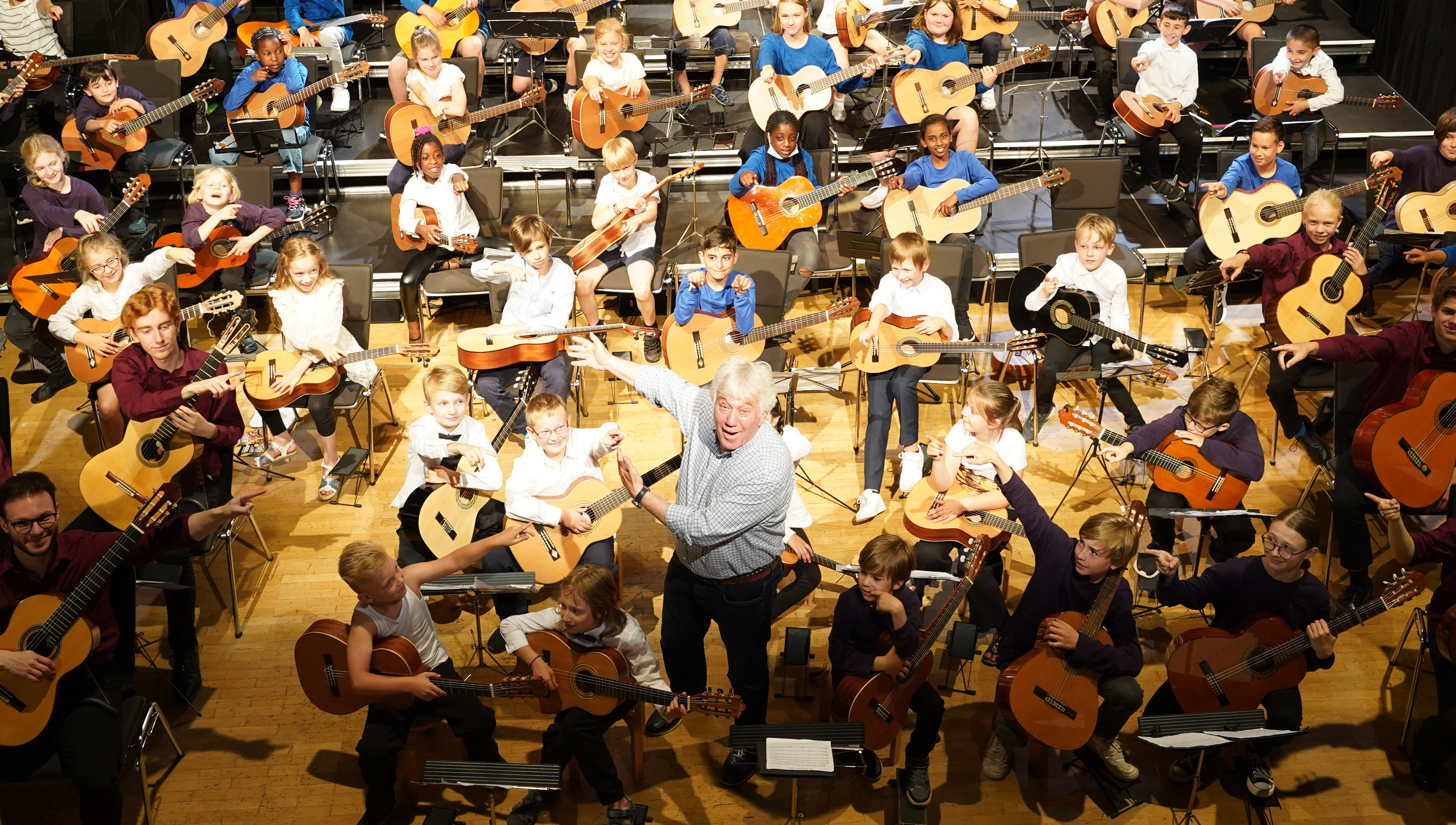 Viele Kinder mit Gitarren versammeln sich auf einer Bühne; ein Dirigent hebt die Arme, bereit für eine Musikaufführung.