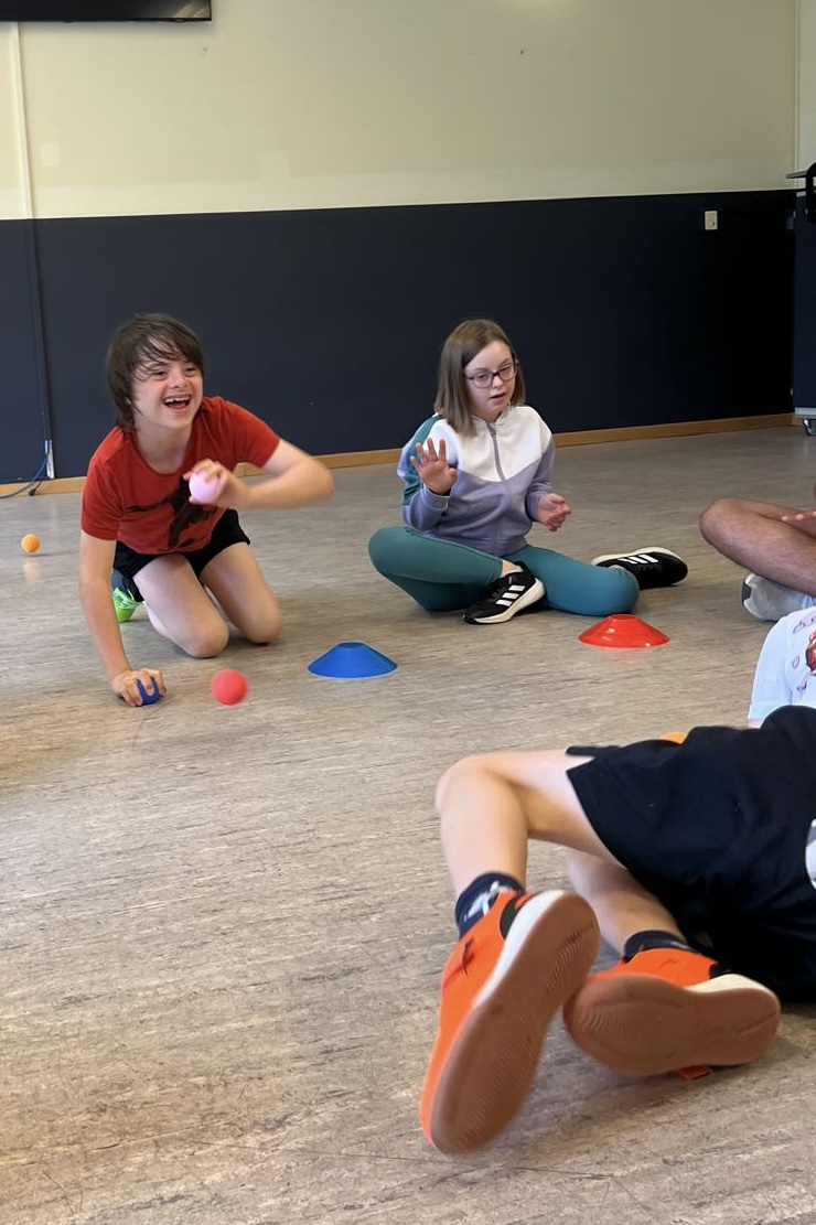 Zwei Kinder spielen in einer Sporthalle auf dem Boden mit bunten Hütchen und Bällen; der Junge in rotem Shirt lacht.