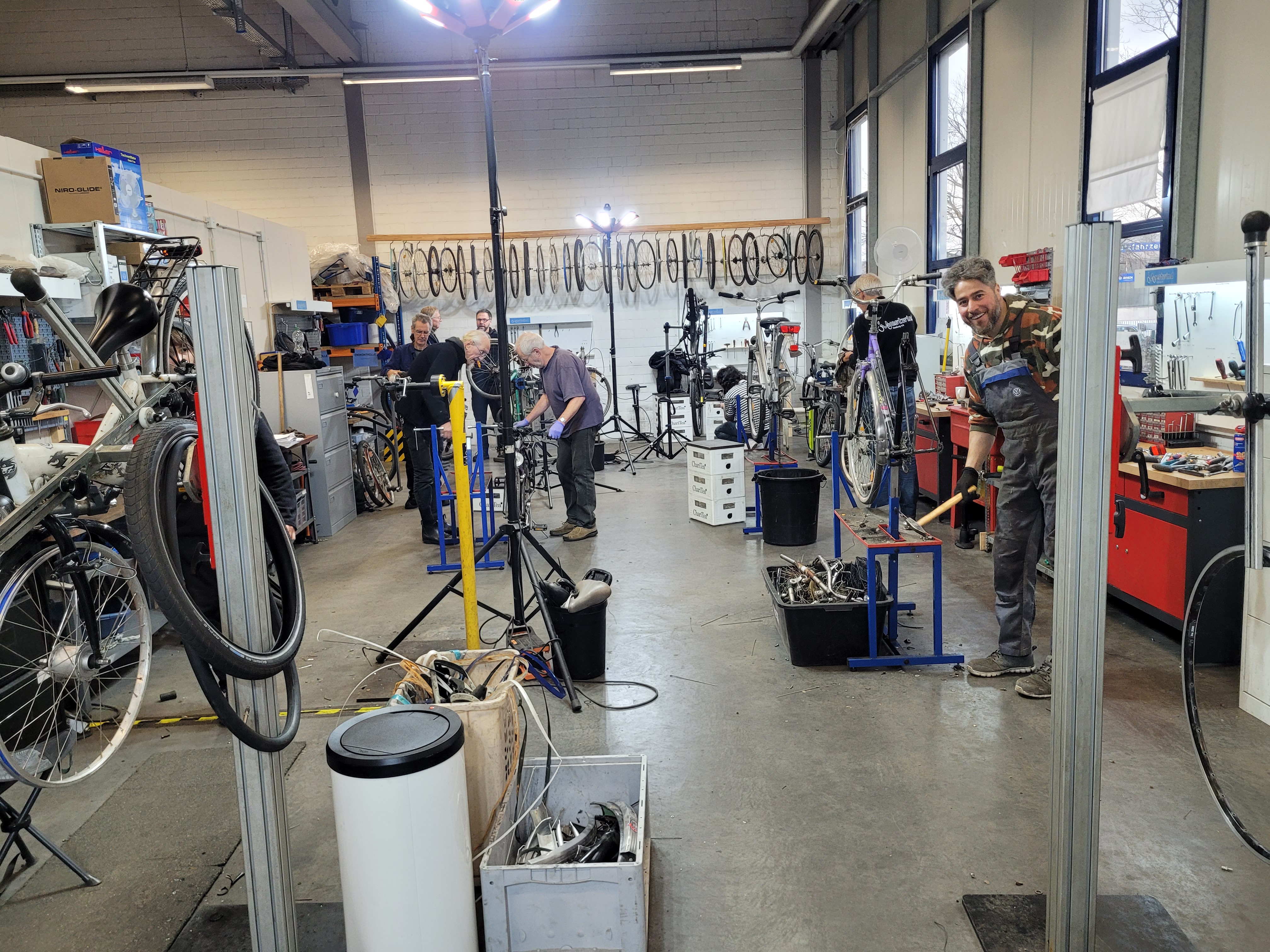 Belebte Fahrradwerkstatt: mehrere Techniker arbeiten an Fahrrädern, Wände voll Laufräder, Werkzeuge und Regale.