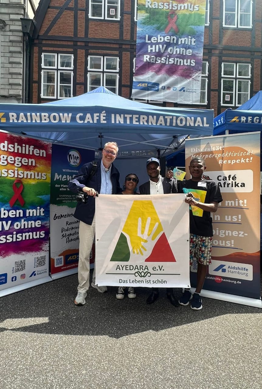 Vier Personen posieren mit AYEDARA e.V.-Banner vor einem Infostand gegen Rassismus.