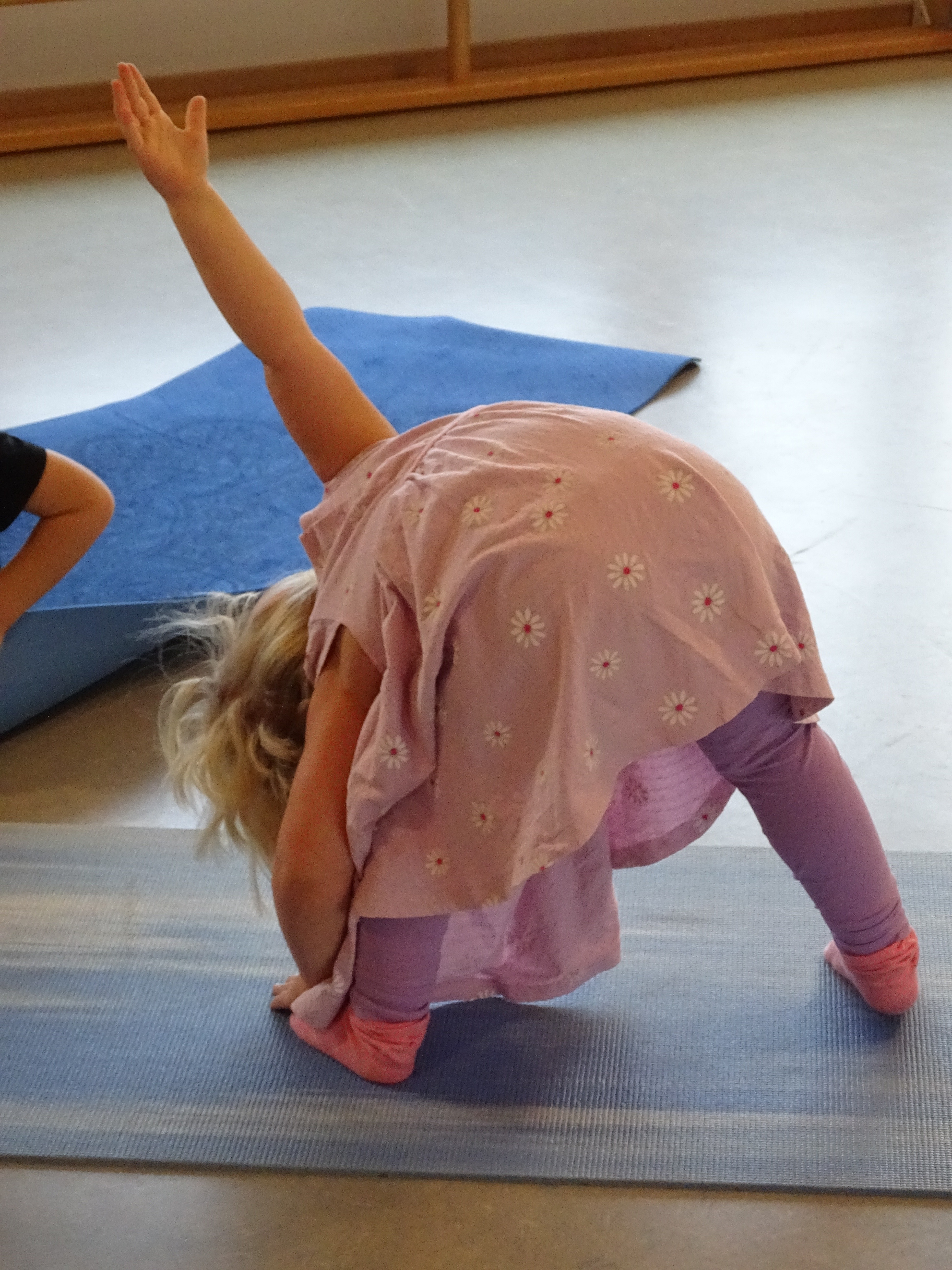 Ein Mädchen in rosa Kleid mit Blumen macht Yoga auf einer blauen Matte; Arm nach oben gestreckt.