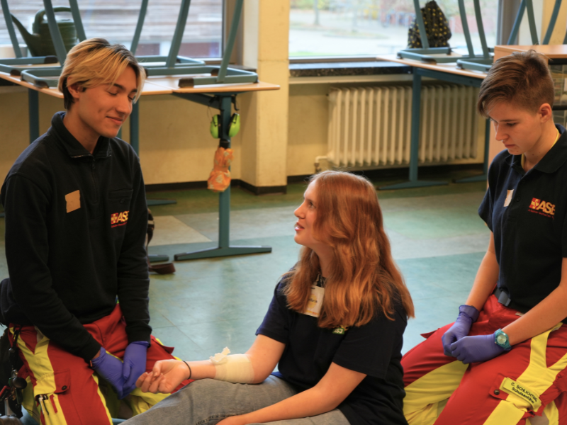 Drei Jugendliche in einer Schule üben Erste Hilfe, eine sitzende Person mit Verband wird betreut.