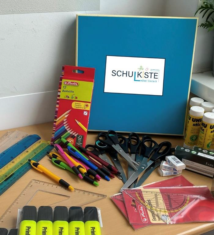 Schulmaterialien auf einem Tisch: Buntstifte, Marker, Lineale, Scheren, Klebestifte und blaues Schild mit Schullogo.
