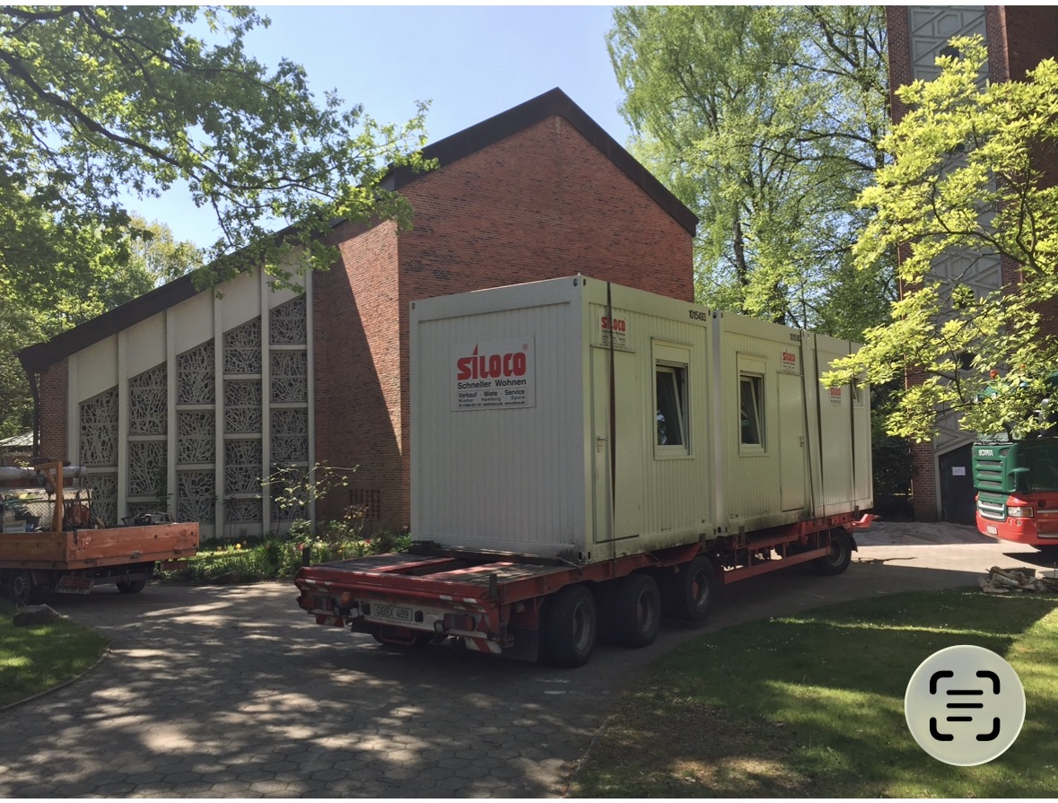 Großer weißer Baucontainer auf einem Mehrachs-Anhänger neben rotem Backsteingebäude und grünen Bäumen.