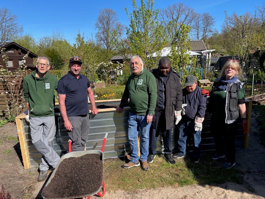 Sechs Personen stehen im Garten hinter einem Metallbehälter; vorne eine Schubkarre mit Erde.