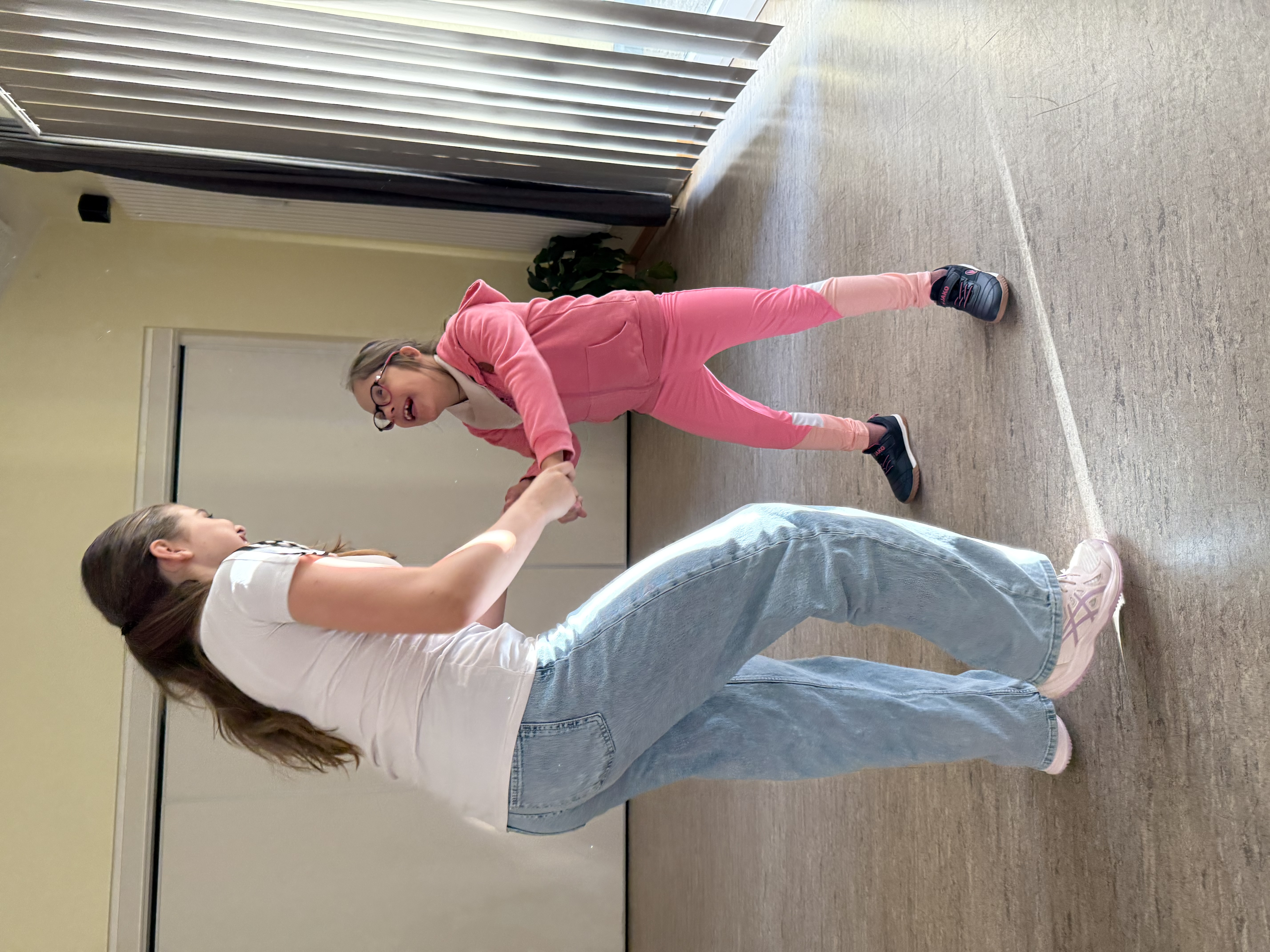 Eine erwachsene Frau hält lachend die Hände eines Mädchens in einem hellen Raum; das Mädchen trägt pinke Kleidung und Brille.