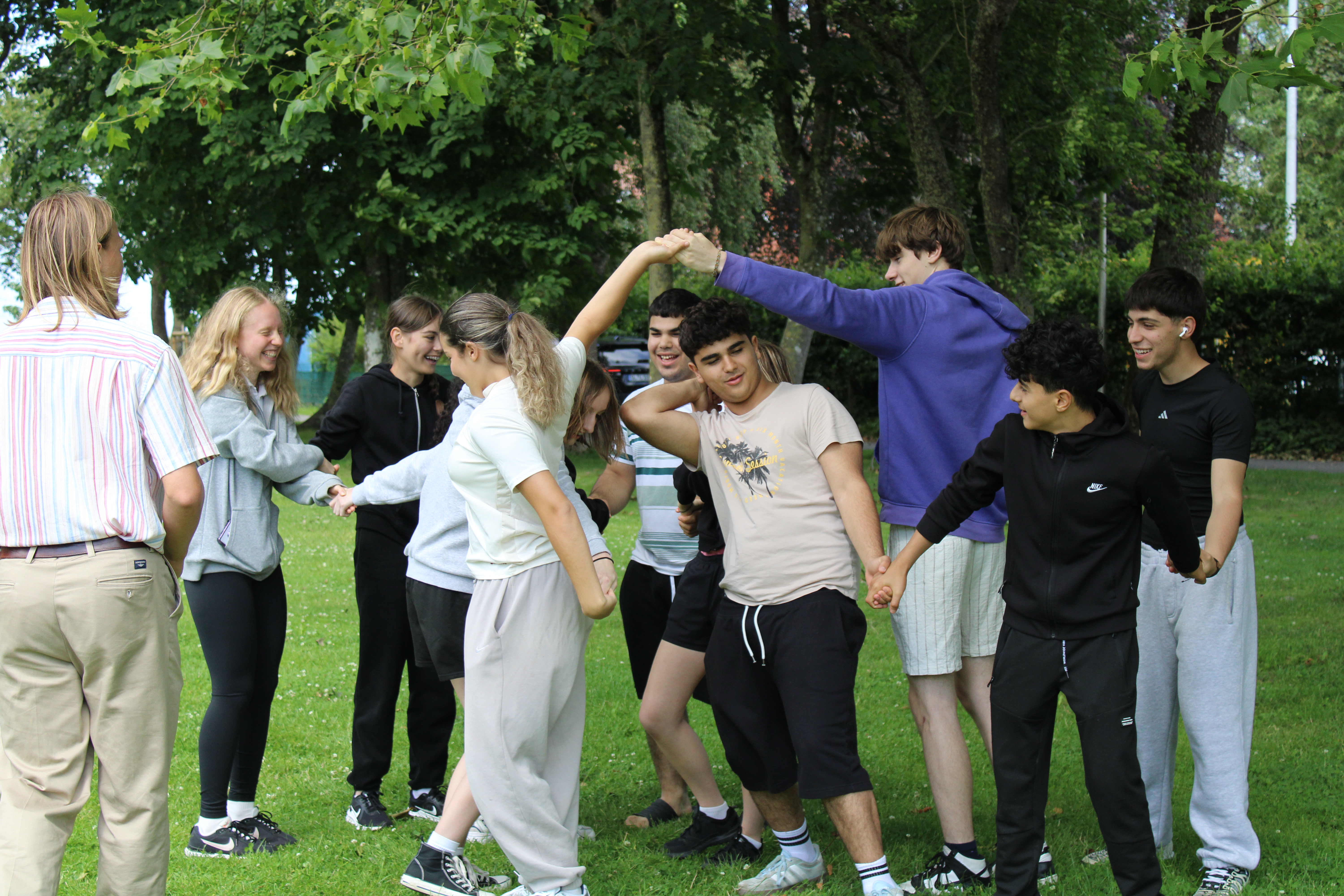 Eine Gruppe Jugendlicher hält sich an den Händen und lacht im Park unter Bäumen.