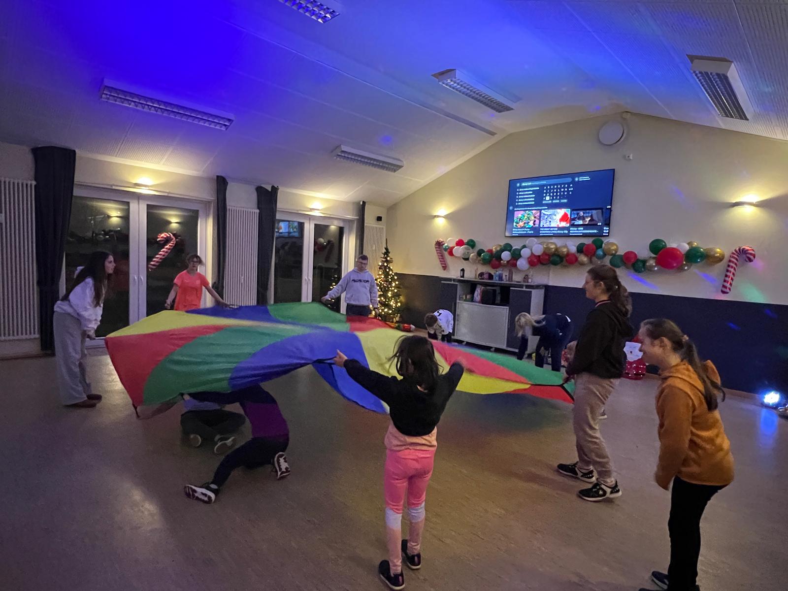 Eine Gruppe Kinder spielt mit einem bunten Regenbogen-Fallschirm in einem festlich geschmückten Raum mit Weihnachtsbaum und Ballons.